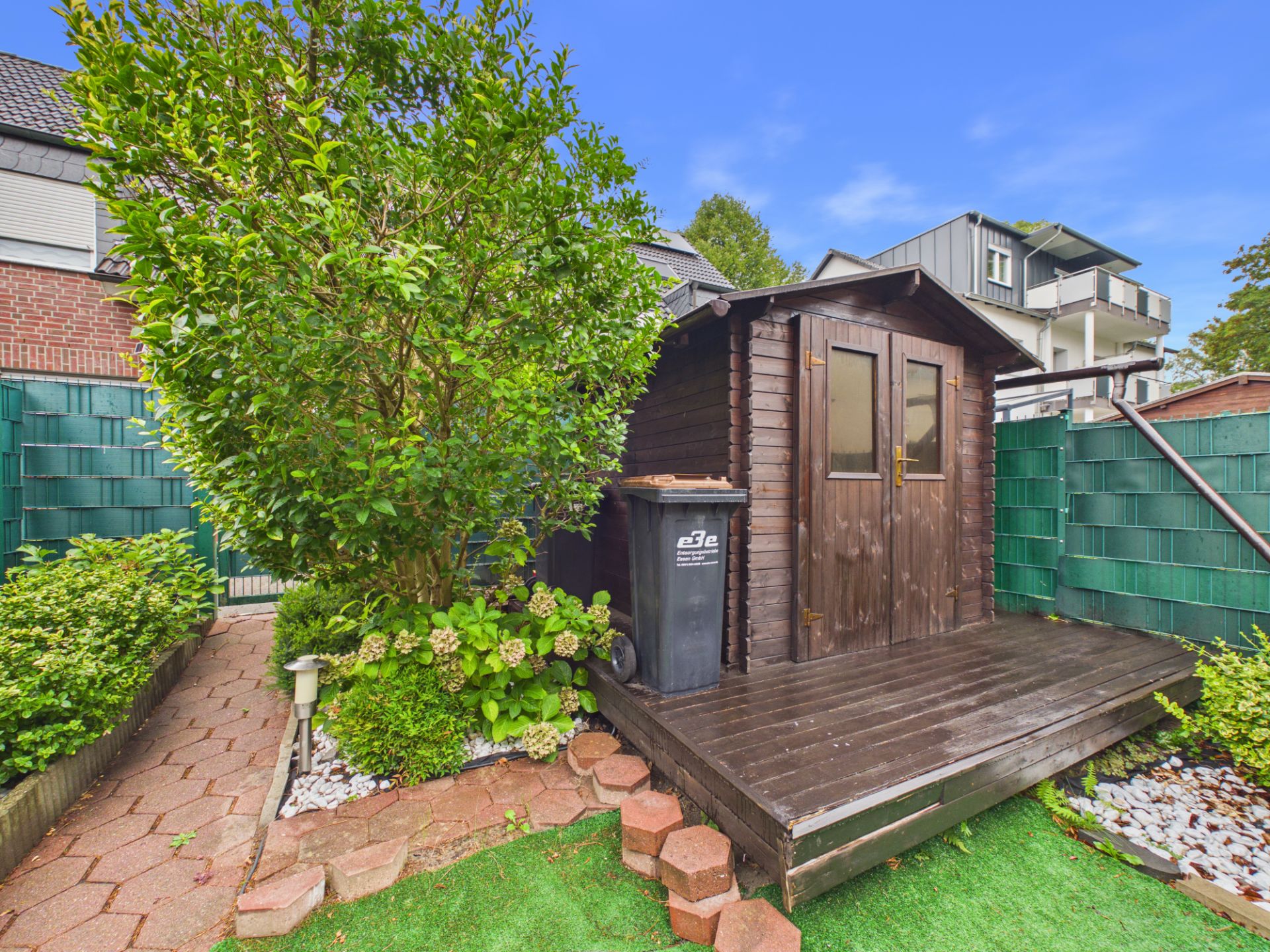 Kleines Holzhaus mit Terrasse im Garten, daneben ein großer Mülltonne und ein Baum.