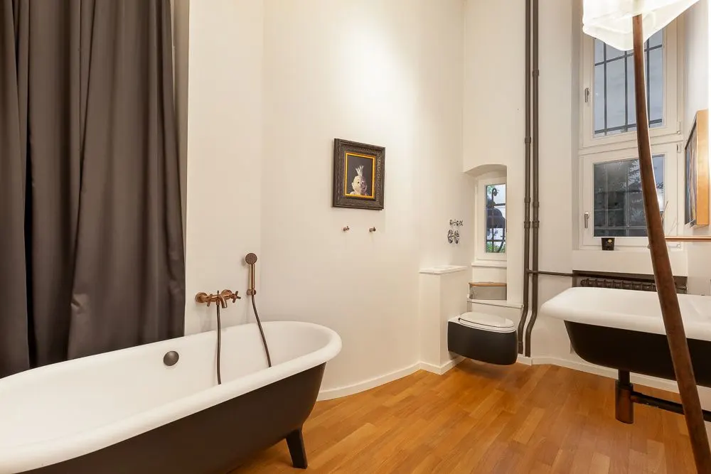 Helles Badezimmer mit freistehender Badewanne, Wand-WC, Holzboden und großem Fenster.