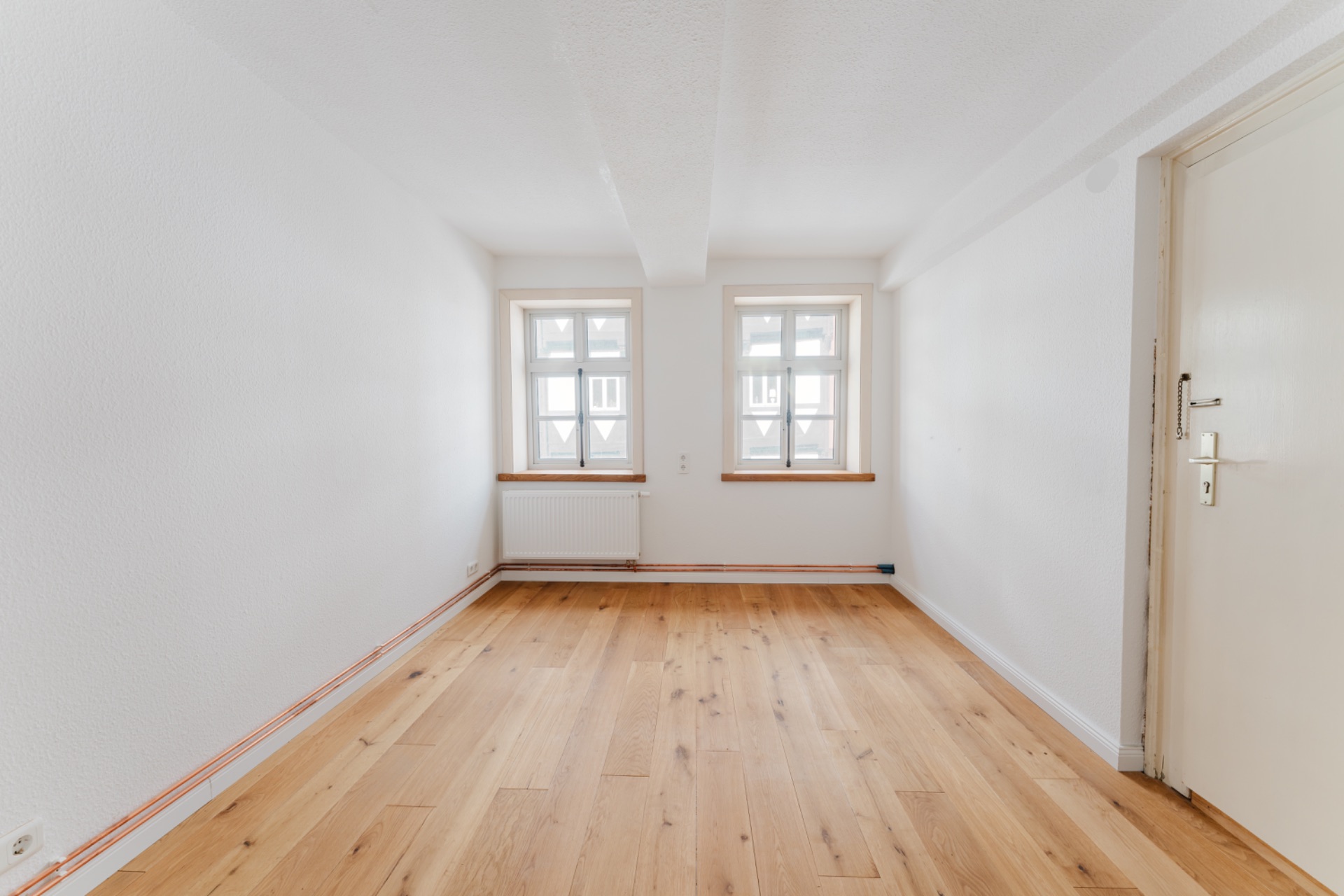 Leerer Raum mit Holzfußboden, weißen Wänden und zwei Fenstern mit Heizung darunter.