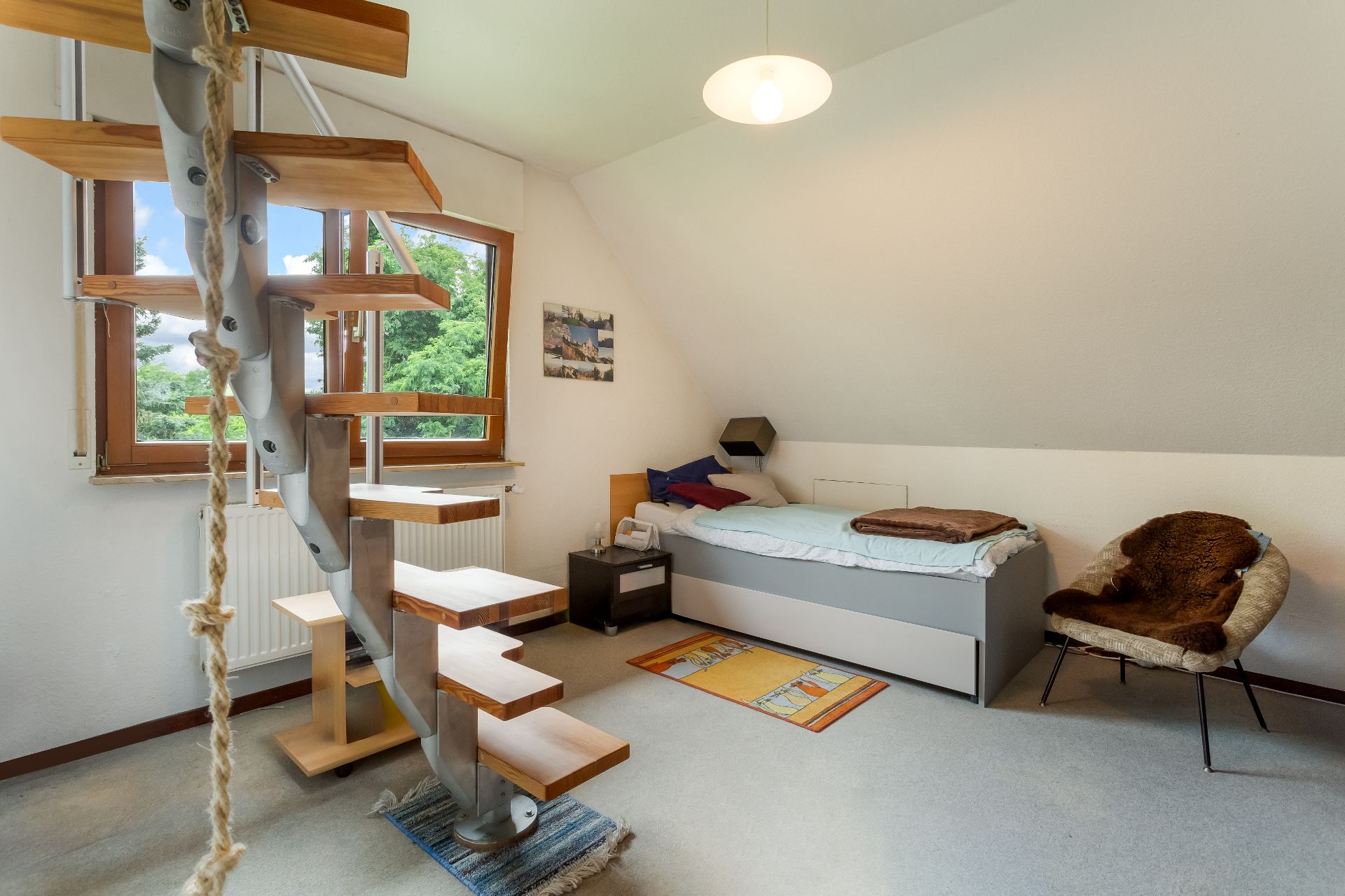 Helles Zimmer mit Einzelbett, Sessel, Wendeltreppe und großem Fenster.