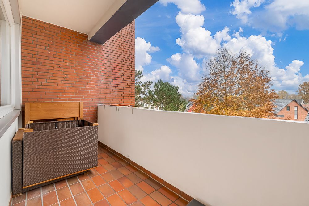 Balkon mit Fliesenboden, Sitztruhe und Ausblick auf Bäume und Häuser.