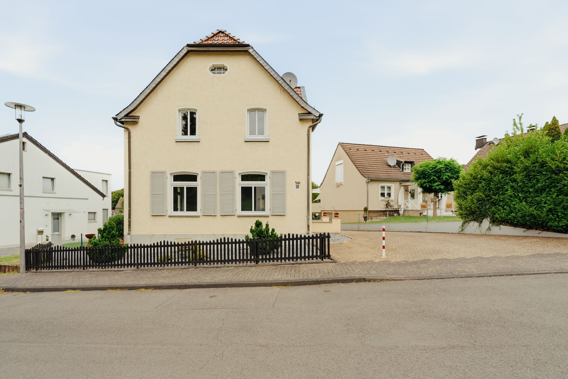 Zweistöckiges Haus mit heller Fassade, Vorgarten und kleinem schwarzen Zaun an einer Straße.