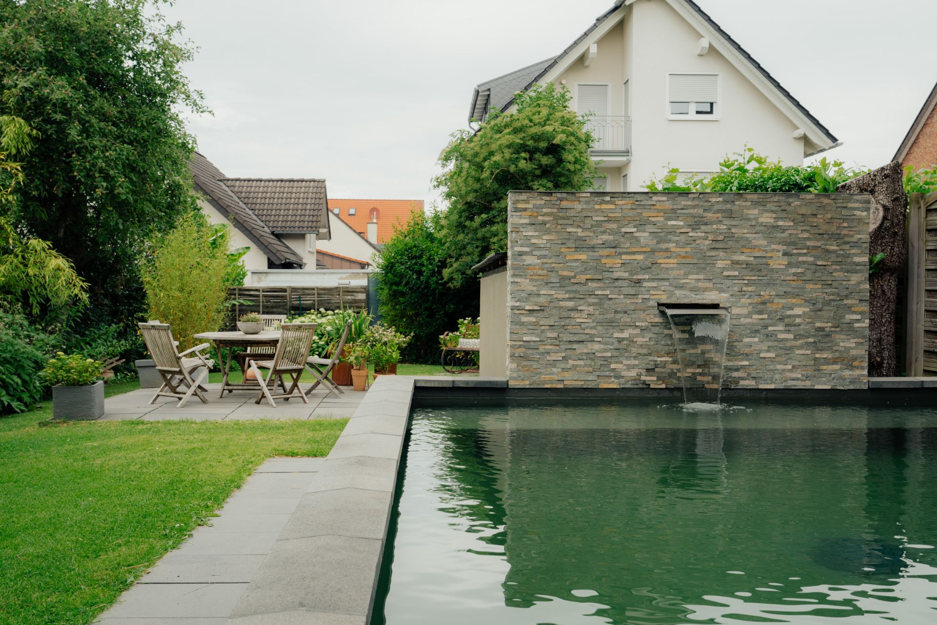Garten mit Pool, Steinwand mit Wasserfall und Sitzbereich mit Tisch und Stühlen.