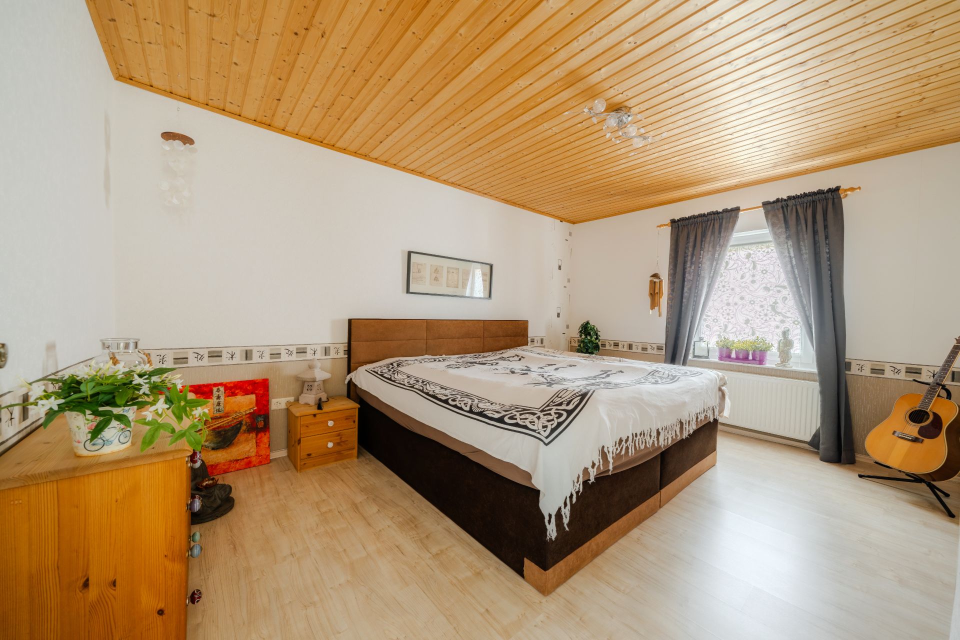 Schlafzimmer mit großem Bett, Holzmöbeln, Gitarre und Fenster mit Vorhängen.