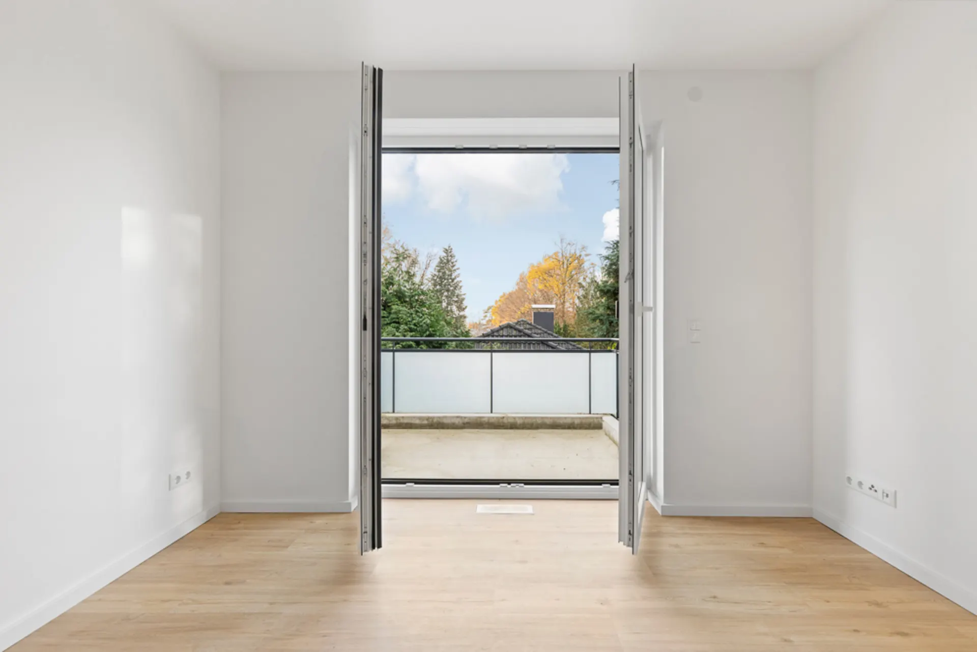 Helles Zimmer mit Holzboden und offener Tür zu einem Balkon mit Blick auf Bäume.