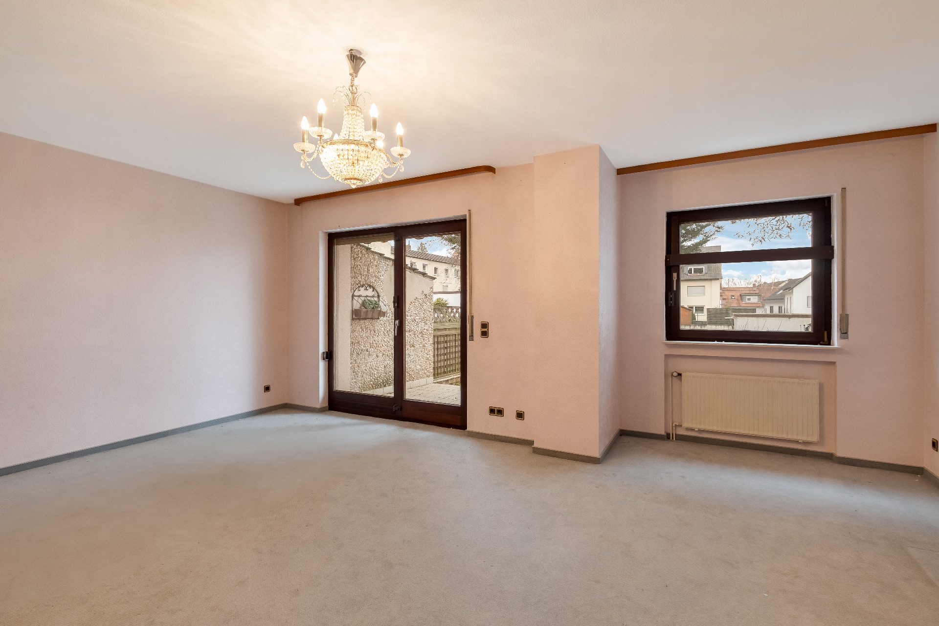 Leeres Wohnzimmer mit Teppich, Kronleuchter, Fenster und Glastür zum Garten.