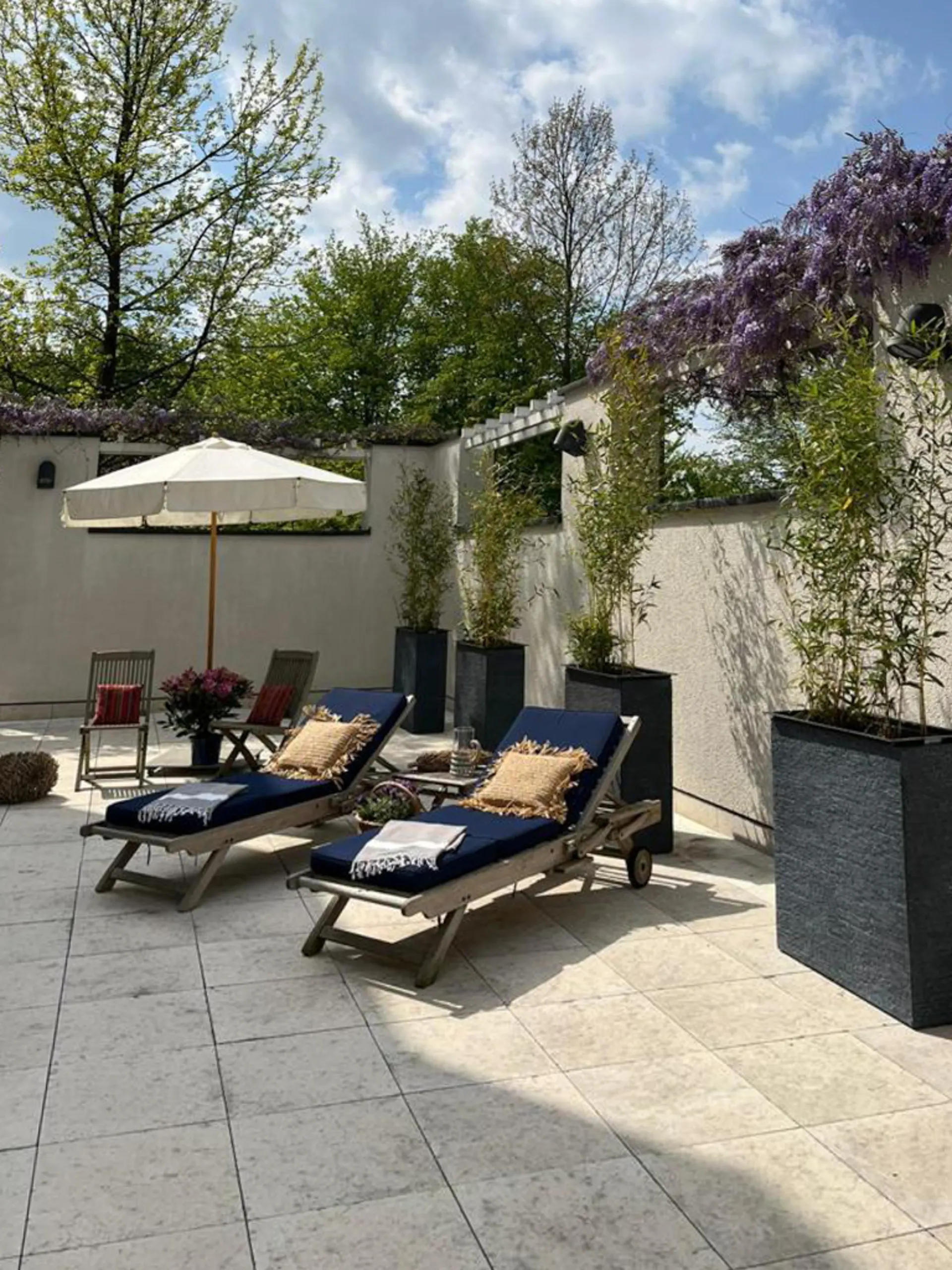 Sonnige Terrasse mit zwei Liegen, Stühlen, Sonnenschirm und großen Pflanzkübeln.