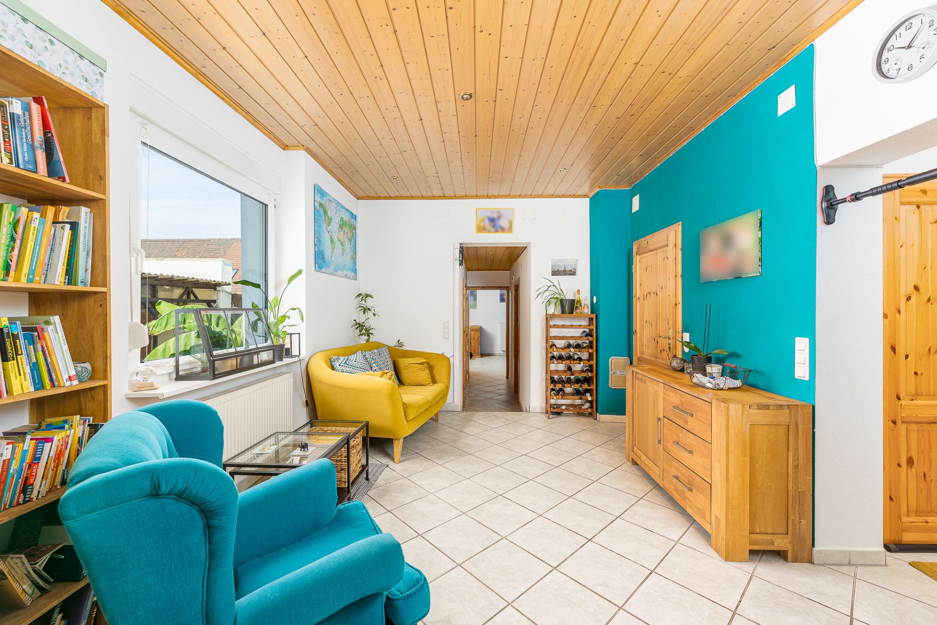 Wohnzimmer mit blauem Sessel, gelbem Sofa, Bücherregal und Holzmöbeln, helle Fliesen und Holzdecke.