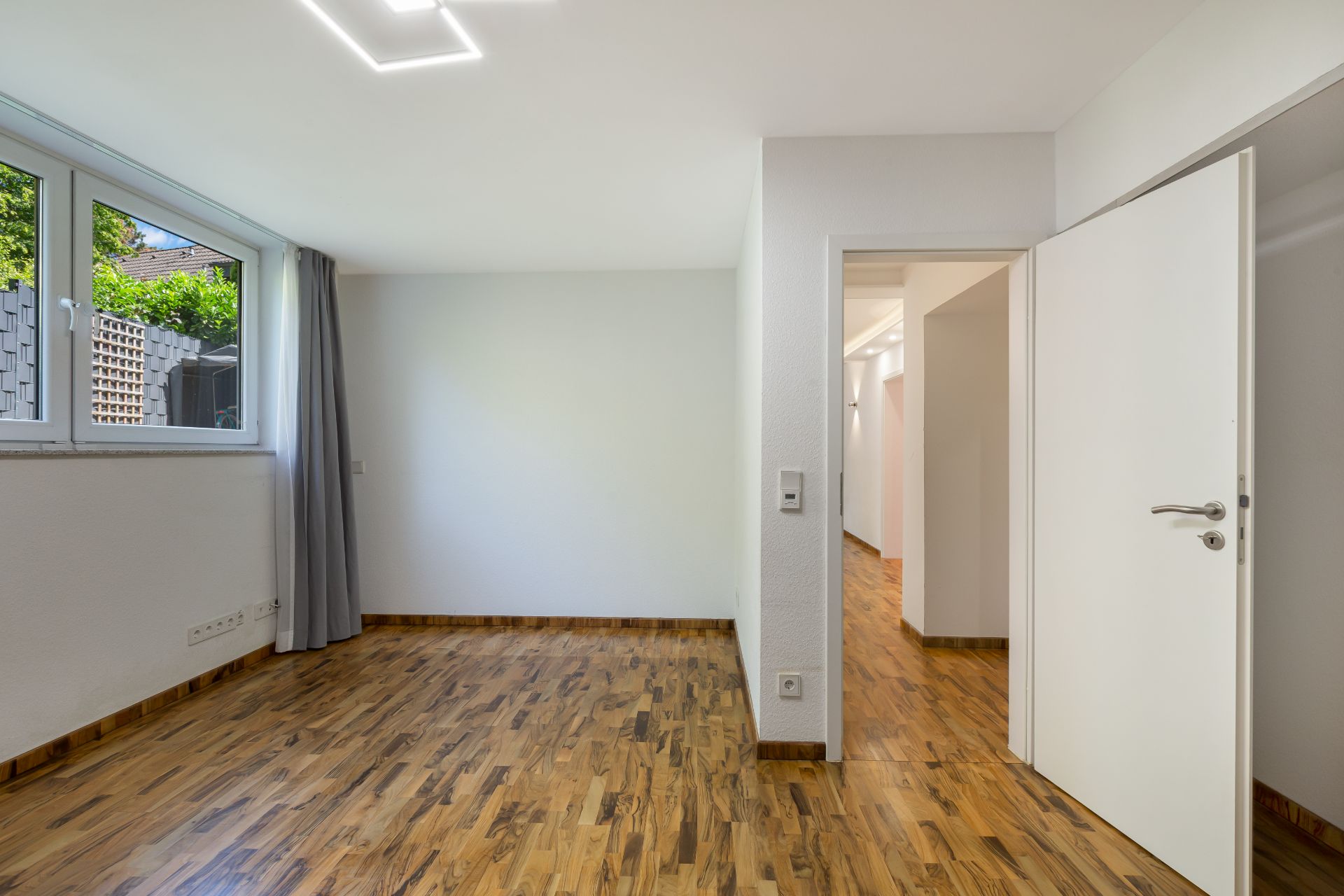 Heller Raum mit Holzfußboden, großem Fenster, weißen Wänden und offen stehender Tür.