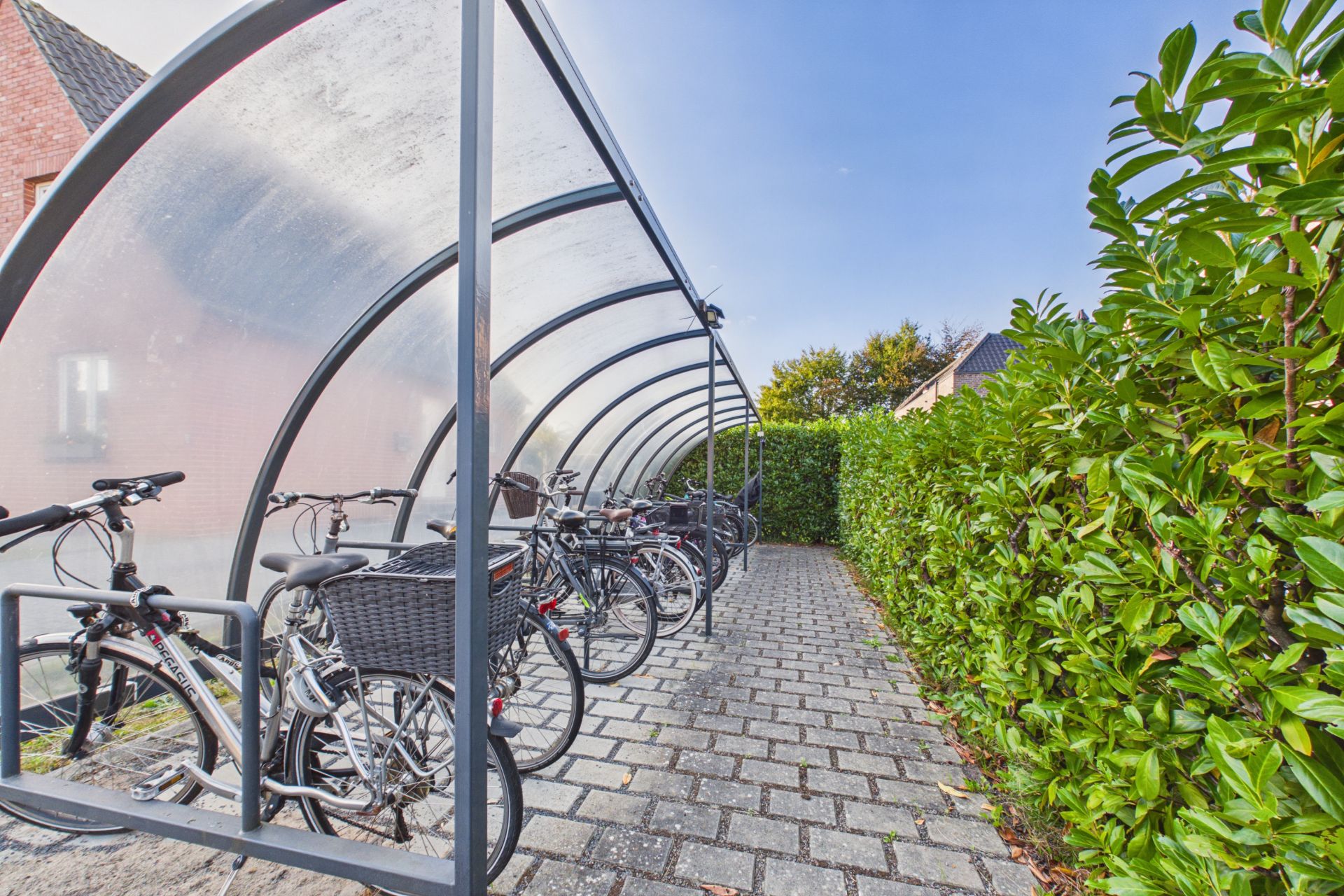 Überdachter Fahrradabstellplatz mit mehreren Fahrrädern auf gepflastertem Weg, umgeben von Hecken.