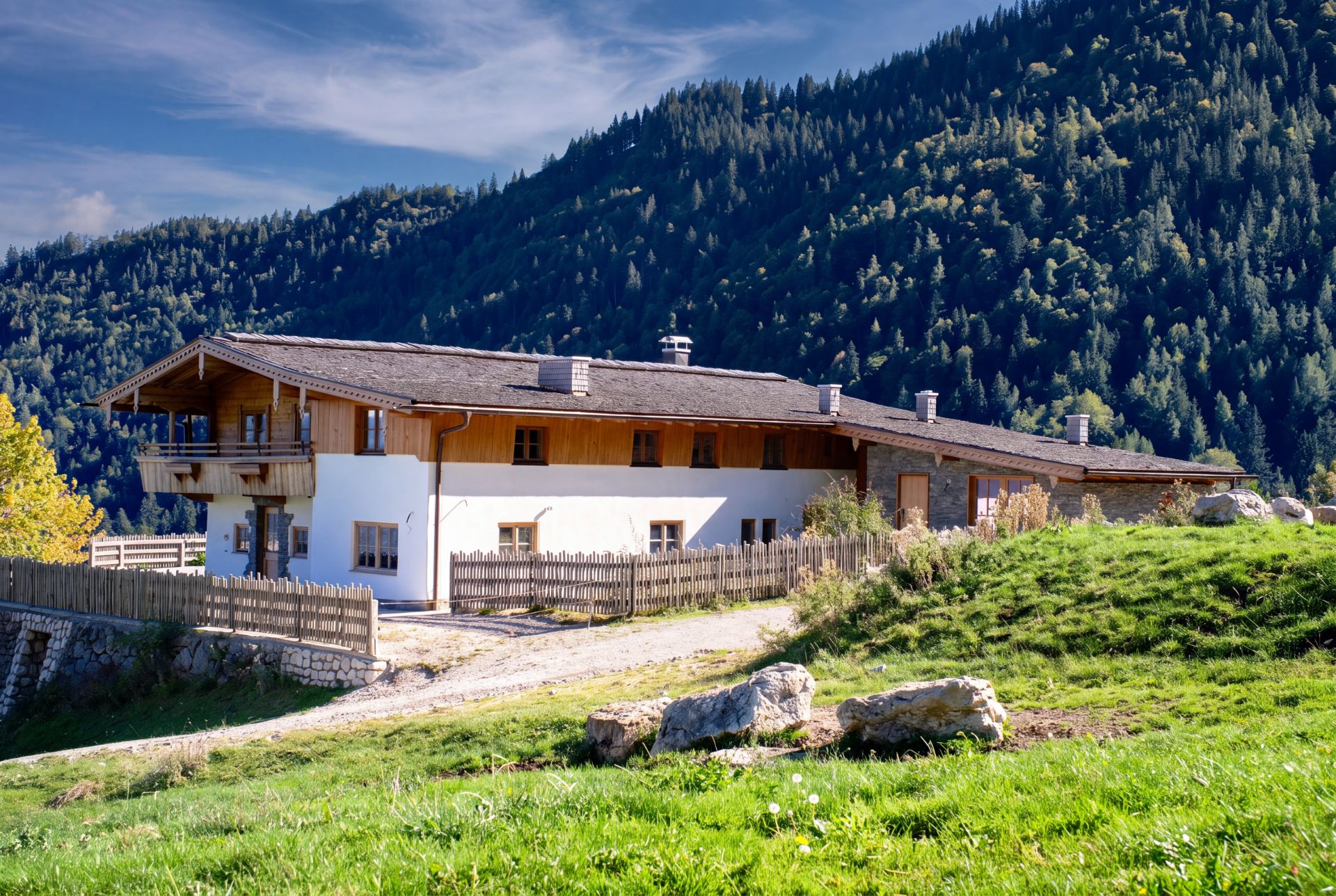 Großes Wohnhaus mit Holzbalkon und Garten, umgeben von Bergen und einem Holzzaun.