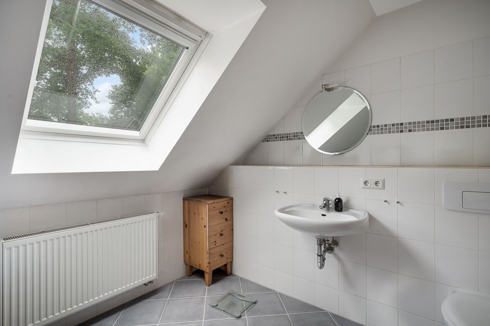 Helles Badezimmer mit Dachfenster, Waschbecken, rundem Spiegel und kleinem Holzschrank.