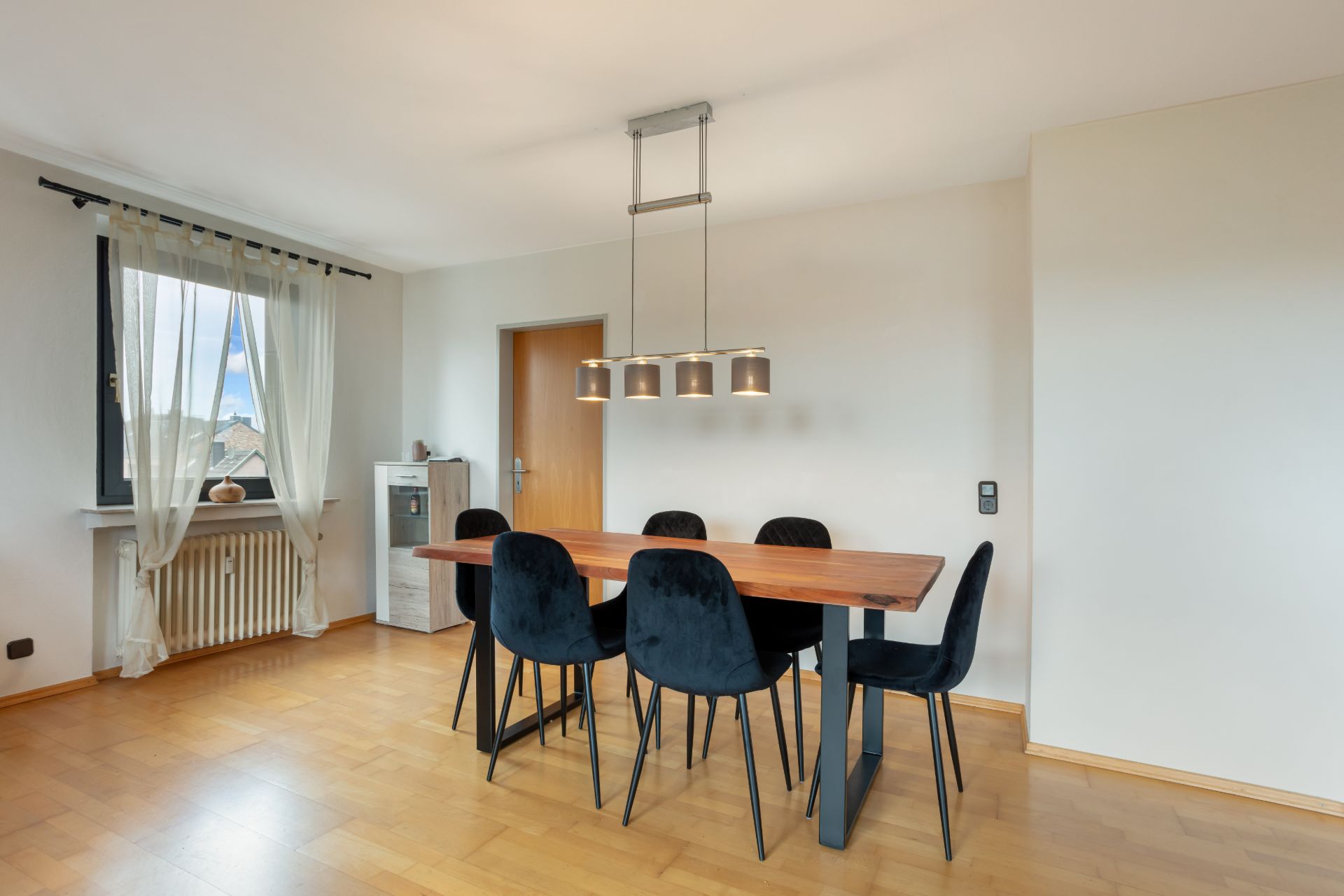 Esszimmer mit Holztisch, sechs schwarzen Stühlen und Deckenlampe, neben Fenster mit Vorhang.