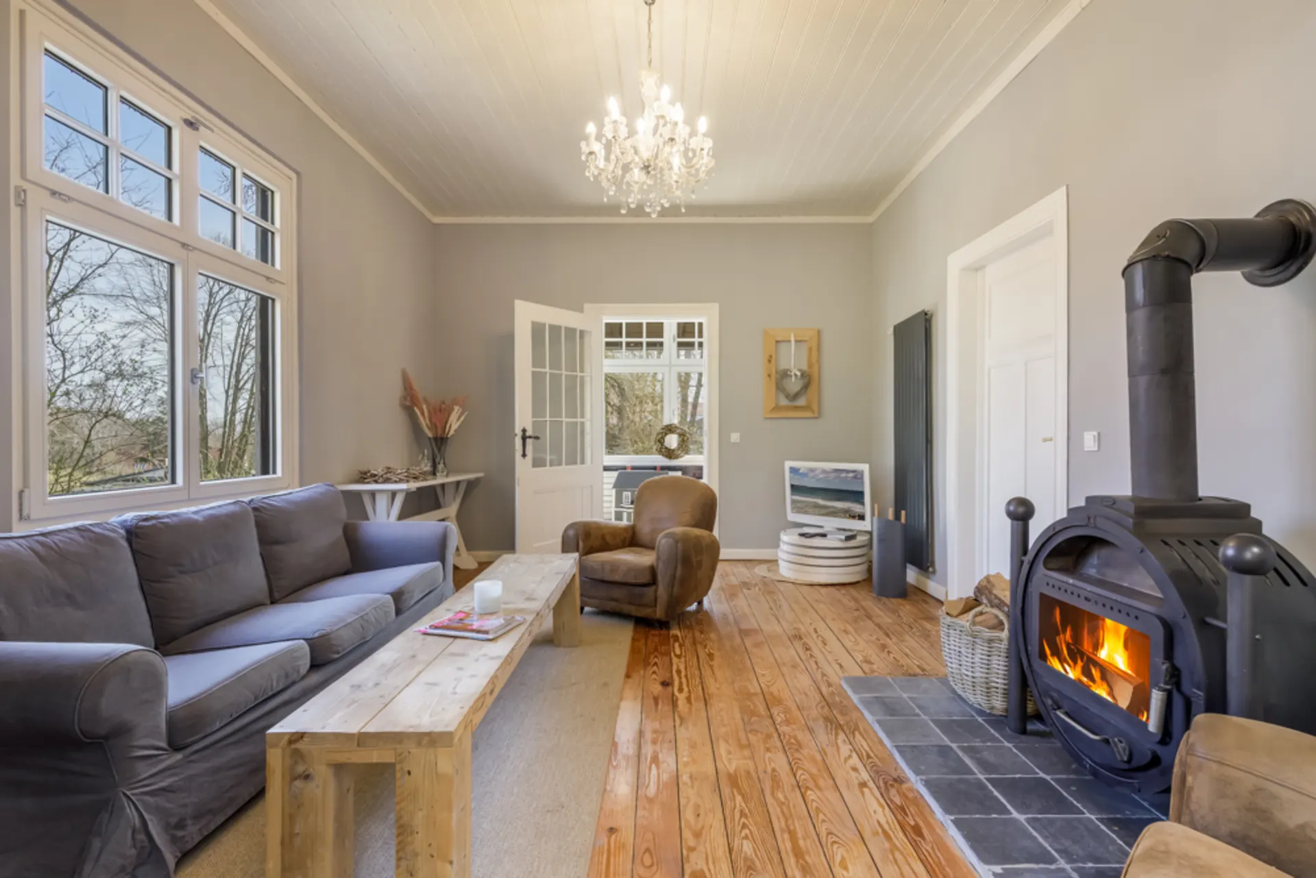 Helles Wohnzimmer mit grauem Sofa, Kaminofen, Holztisch und Kronleuchter.