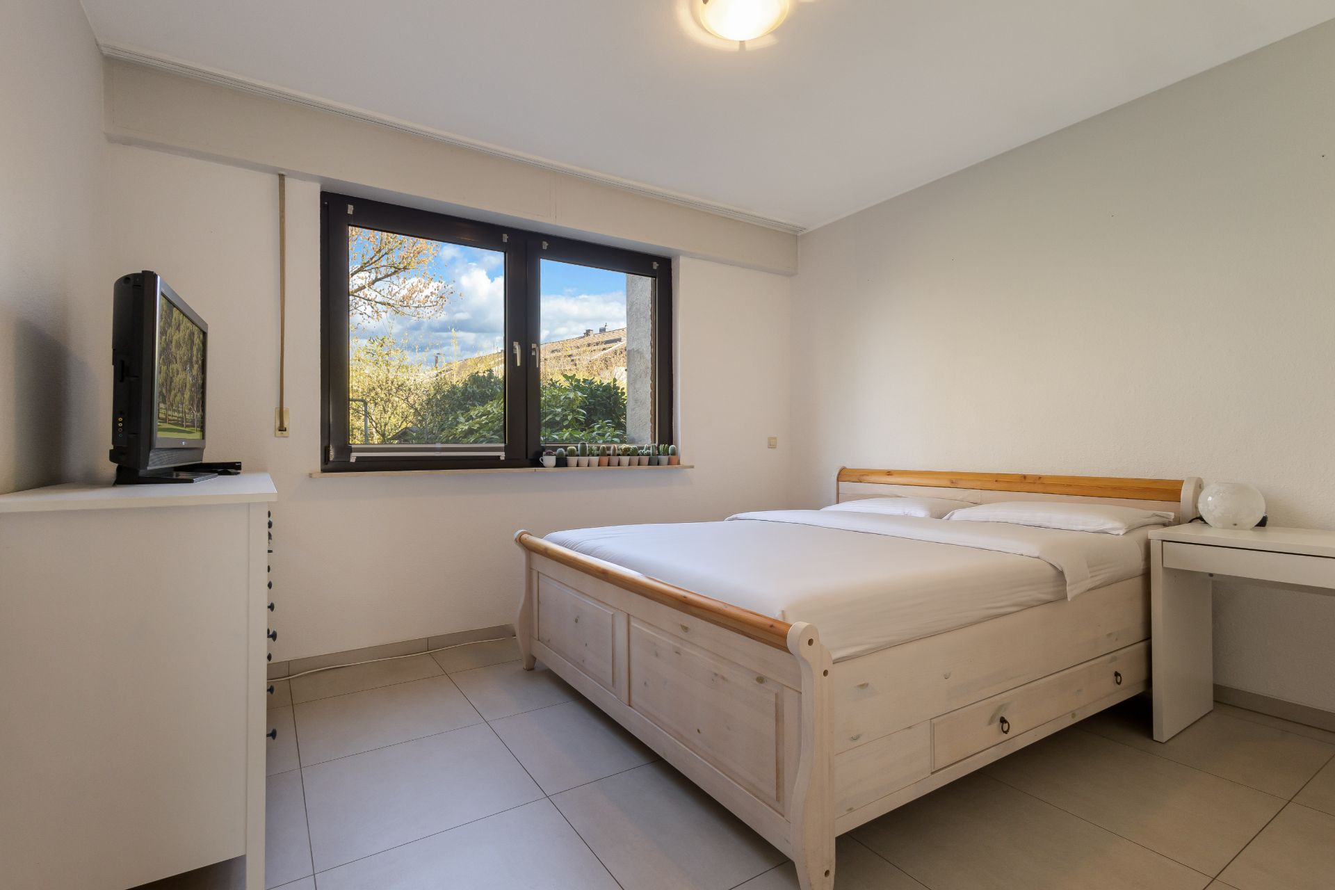 Helles Schlafzimmer mit Doppelbett, Fernseher auf Kommode und großem Fenster mit Ausblick.