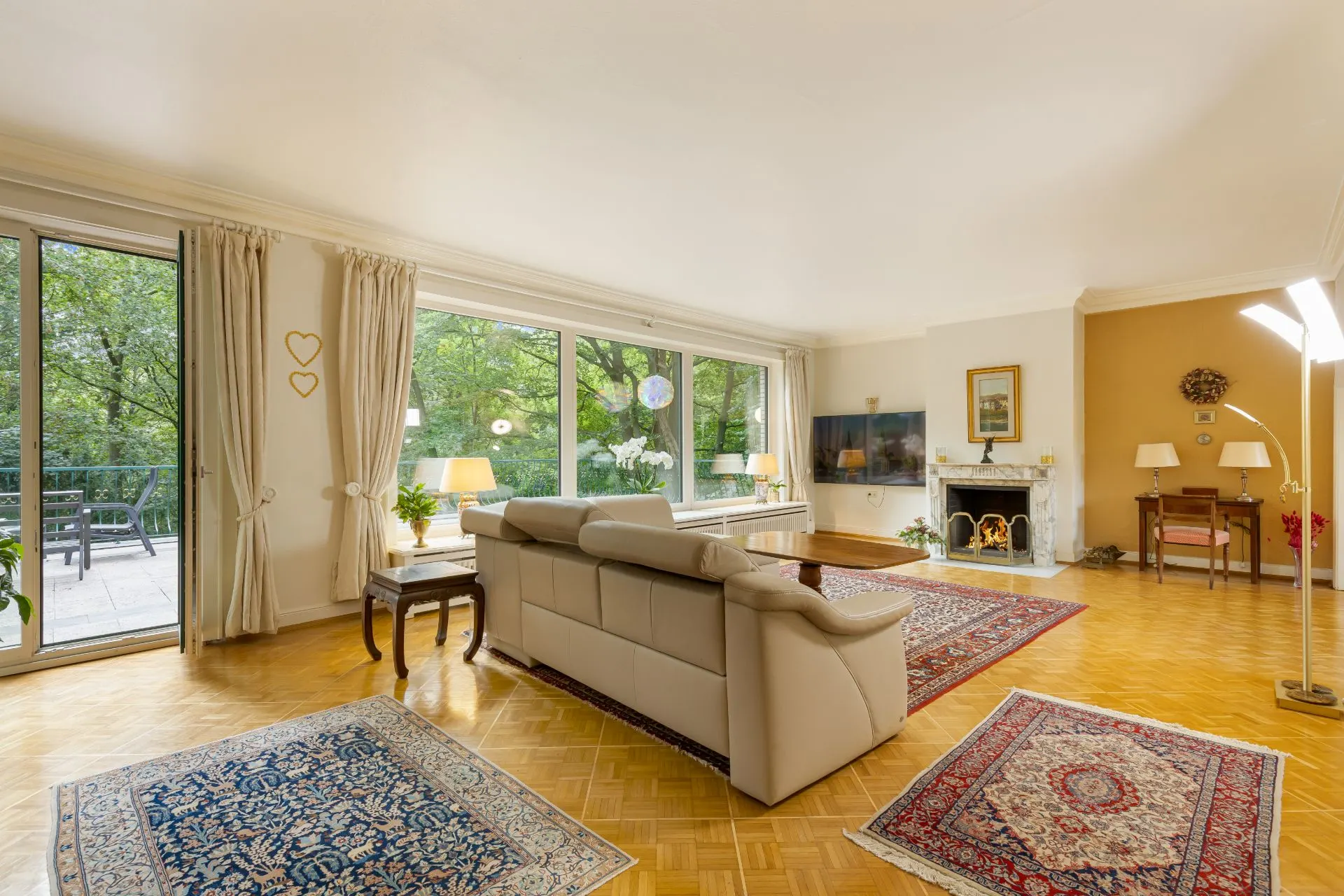 Helles Wohnzimmer mit Sofa, Teppichen, Kamin und großen Fenstern mit Blick ins Grüne.