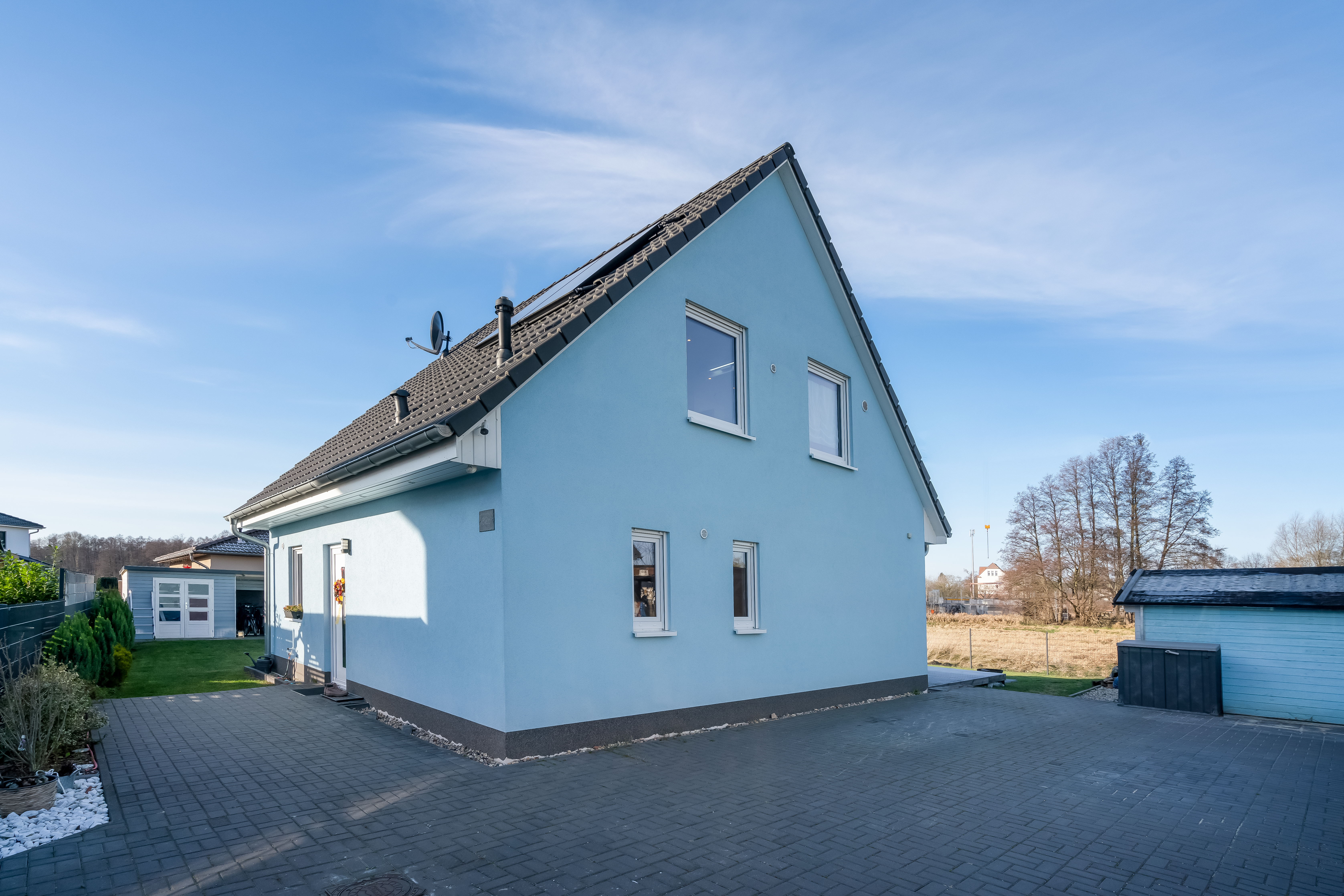 Blaues Einfamilienhaus mit dunklem Dach, gepflastertem Hof und Garten im Hintergrund.