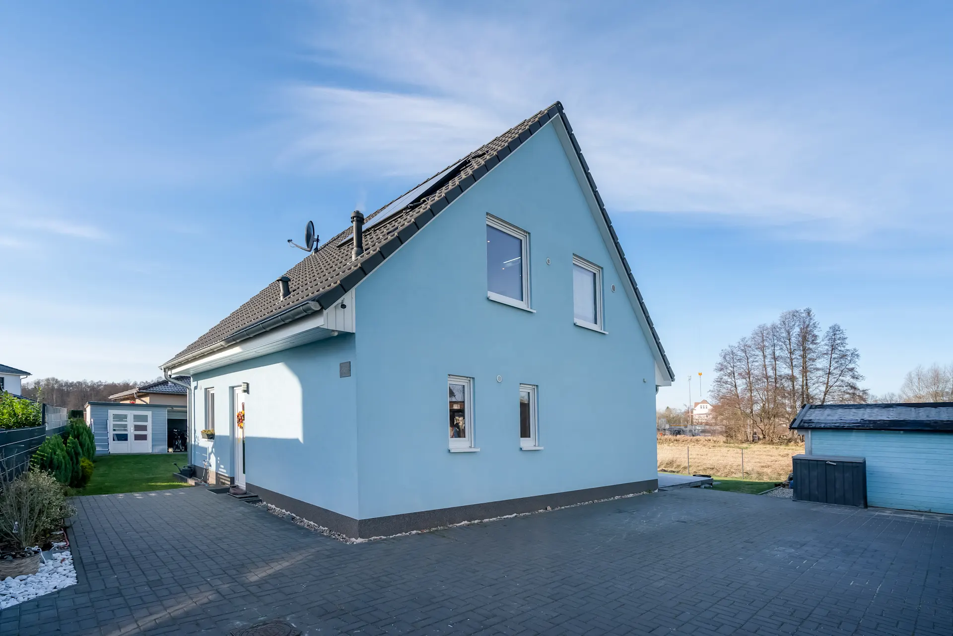 Blaues Einfamilienhaus mit dunklem Dach, gepflastertem Hof und Garten im Hintergrund.