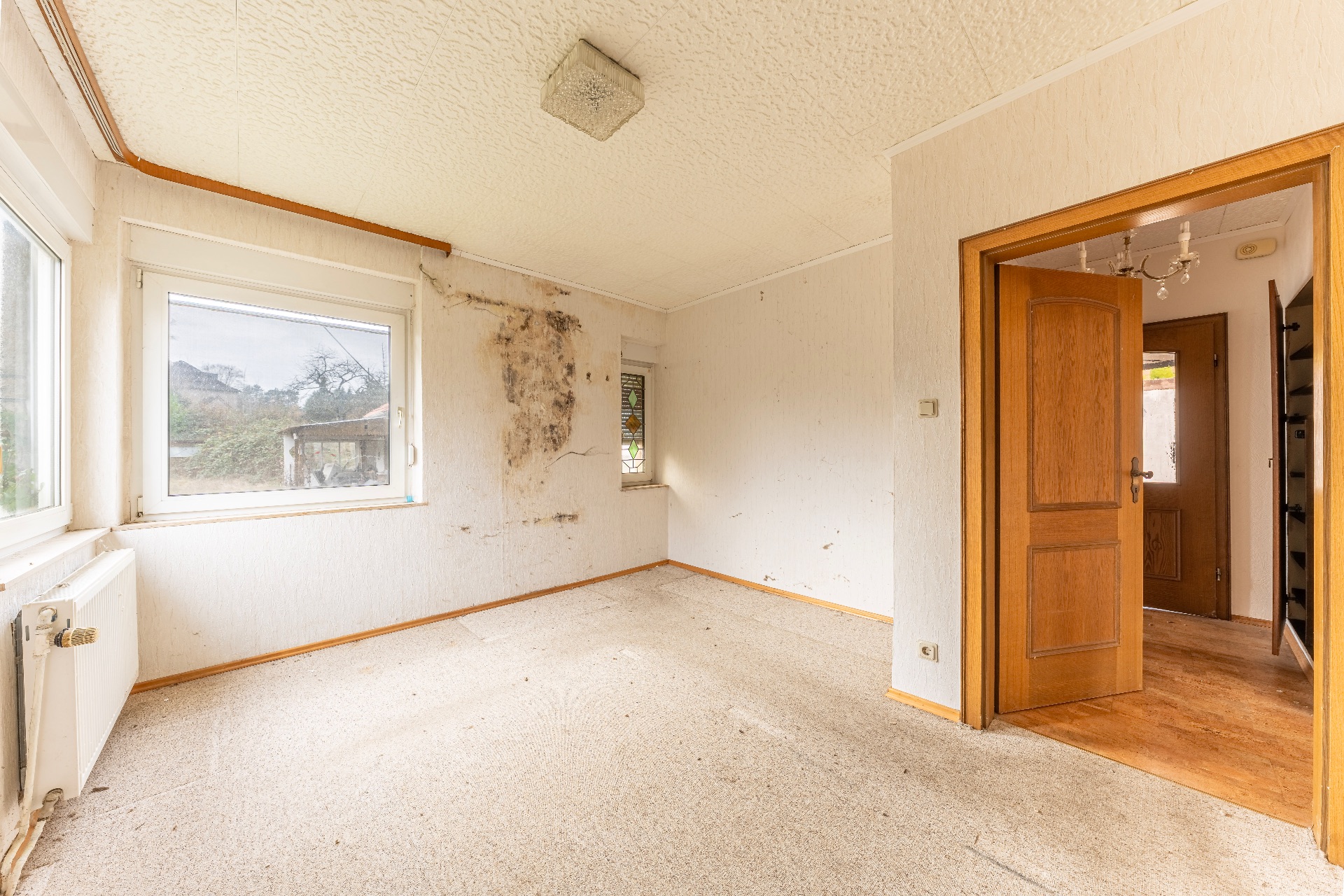 Leeres Zimmer mit zwei Fenstern, sichtbaren Wandschäden und geöffneter Holztür.