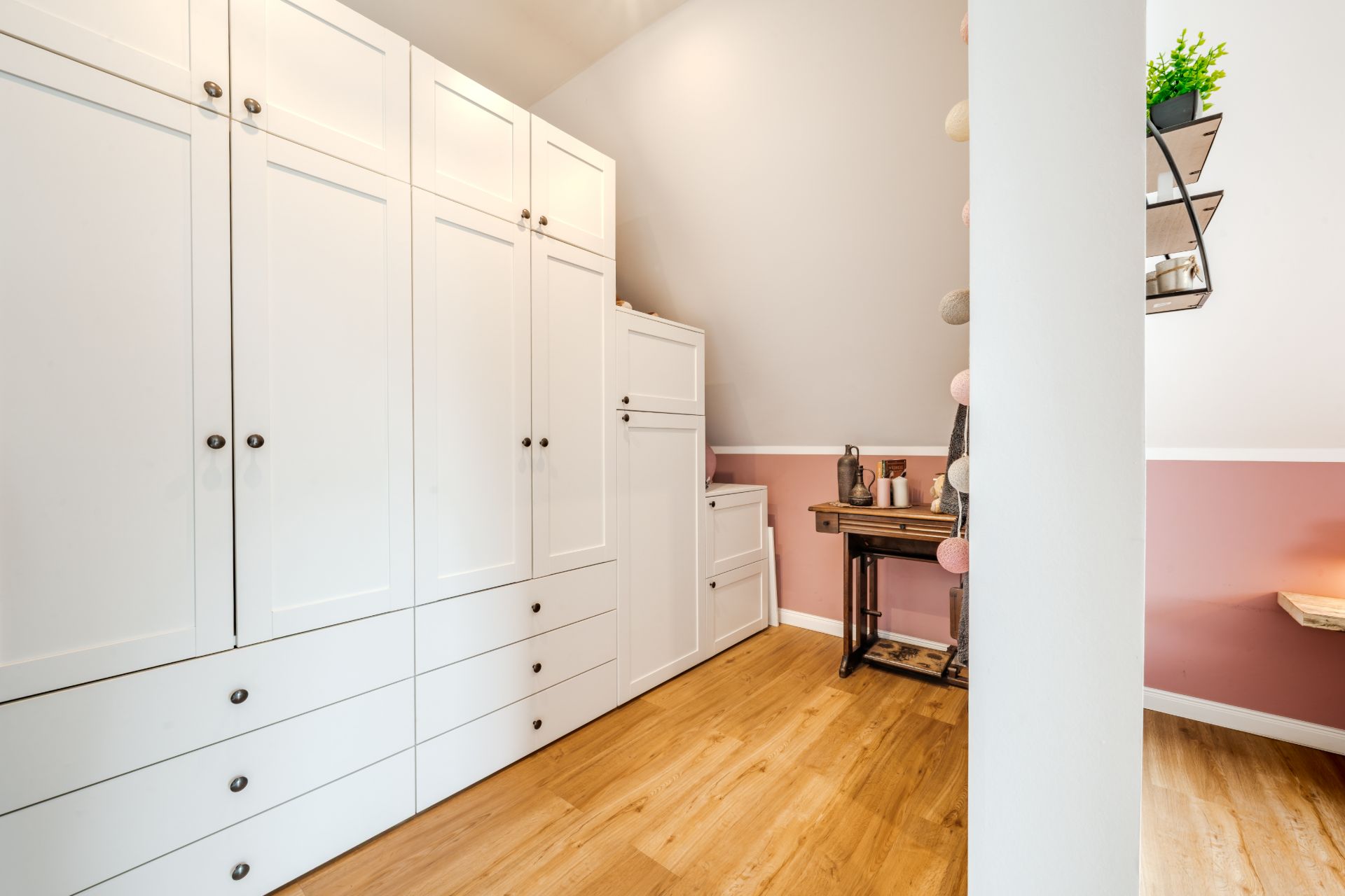 Heller Raum mit weißen Schränken, Holzfußboden und einem kleinen Schreibtisch an einer rosa Wand.