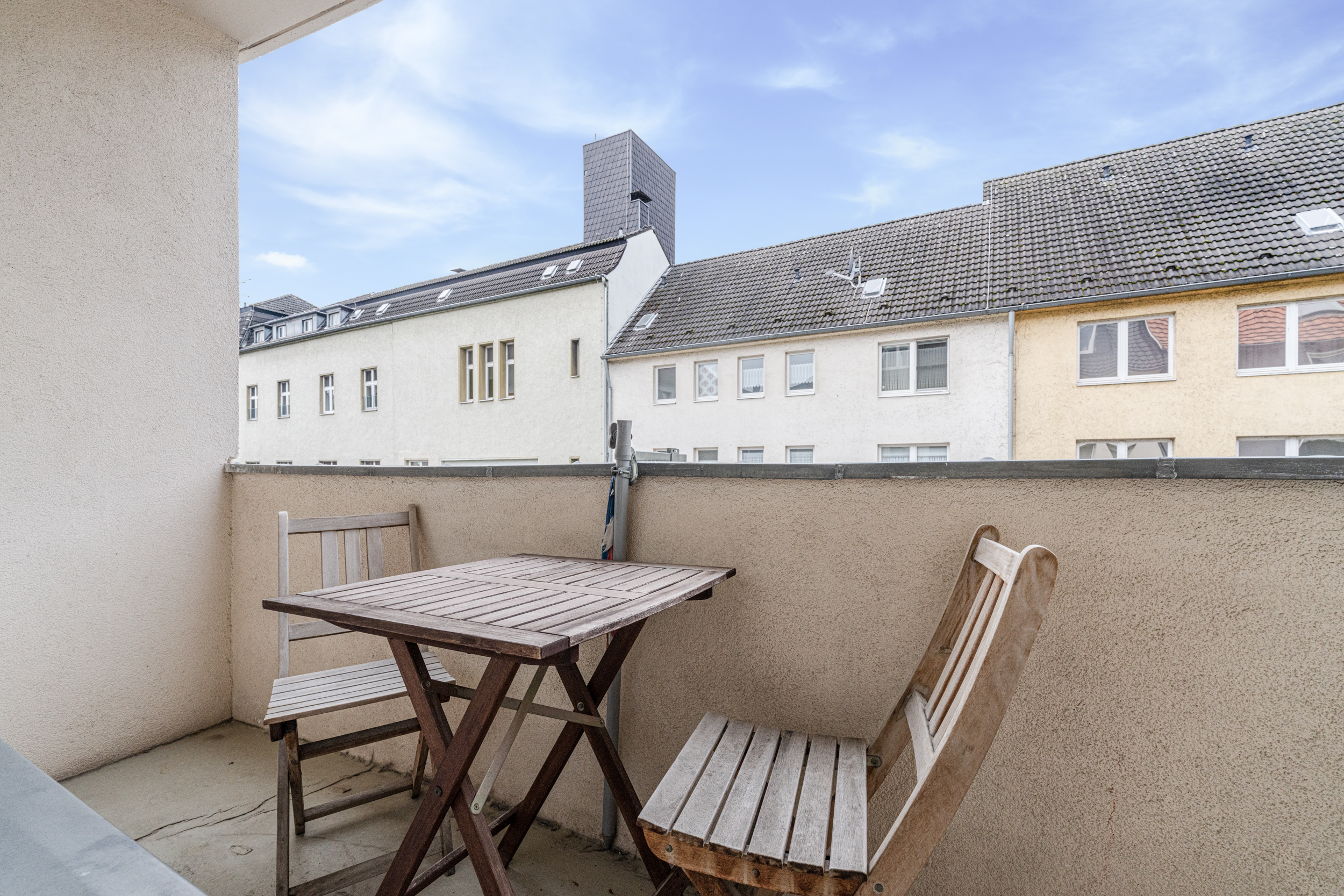Kleiner Balkon mit Holztisch und zwei Holzstühlen, Blick auf Nachbarhäuser.