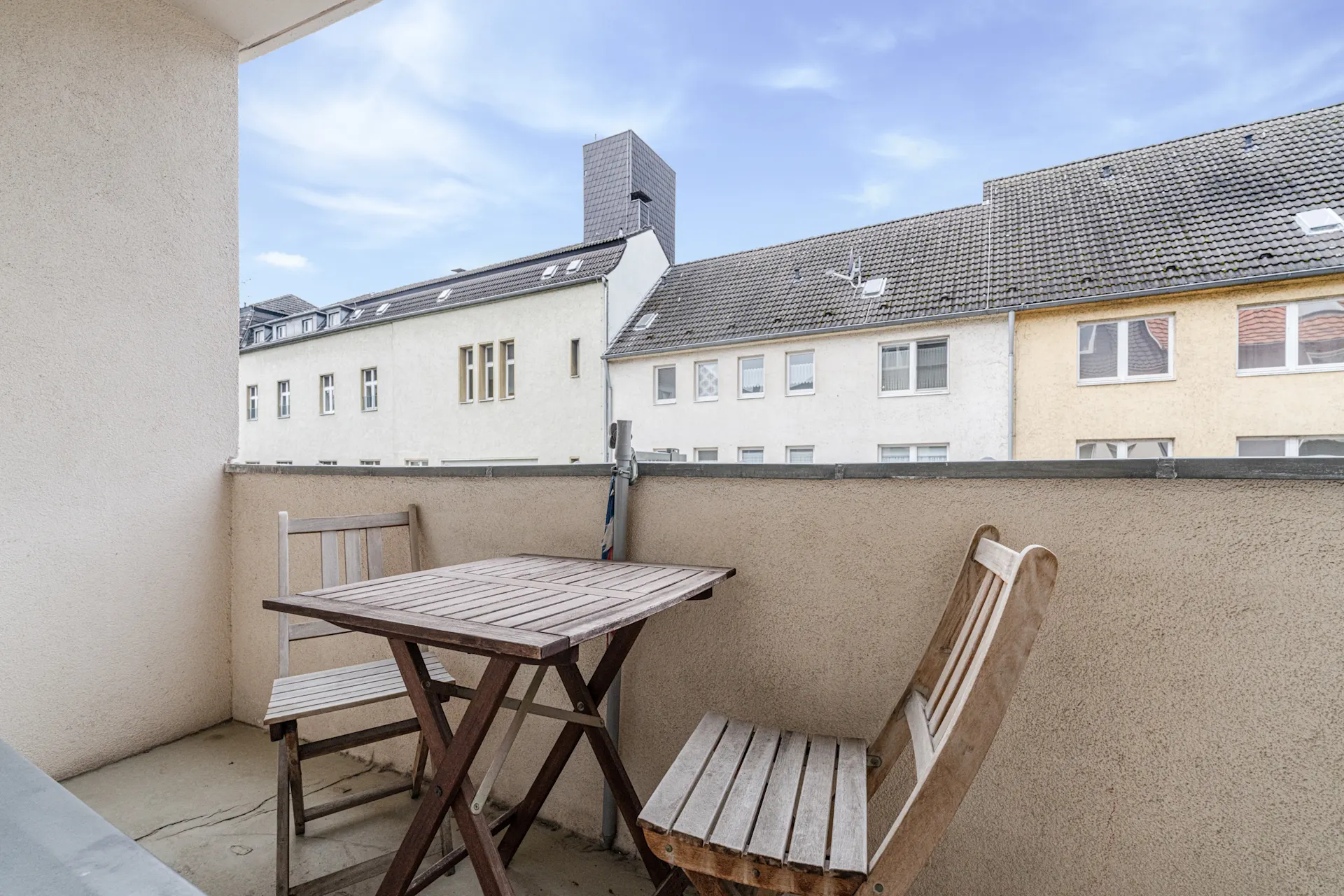 Kleiner Balkon mit Holztisch und zwei Holzstühlen, Blick auf Nachbarhäuser.