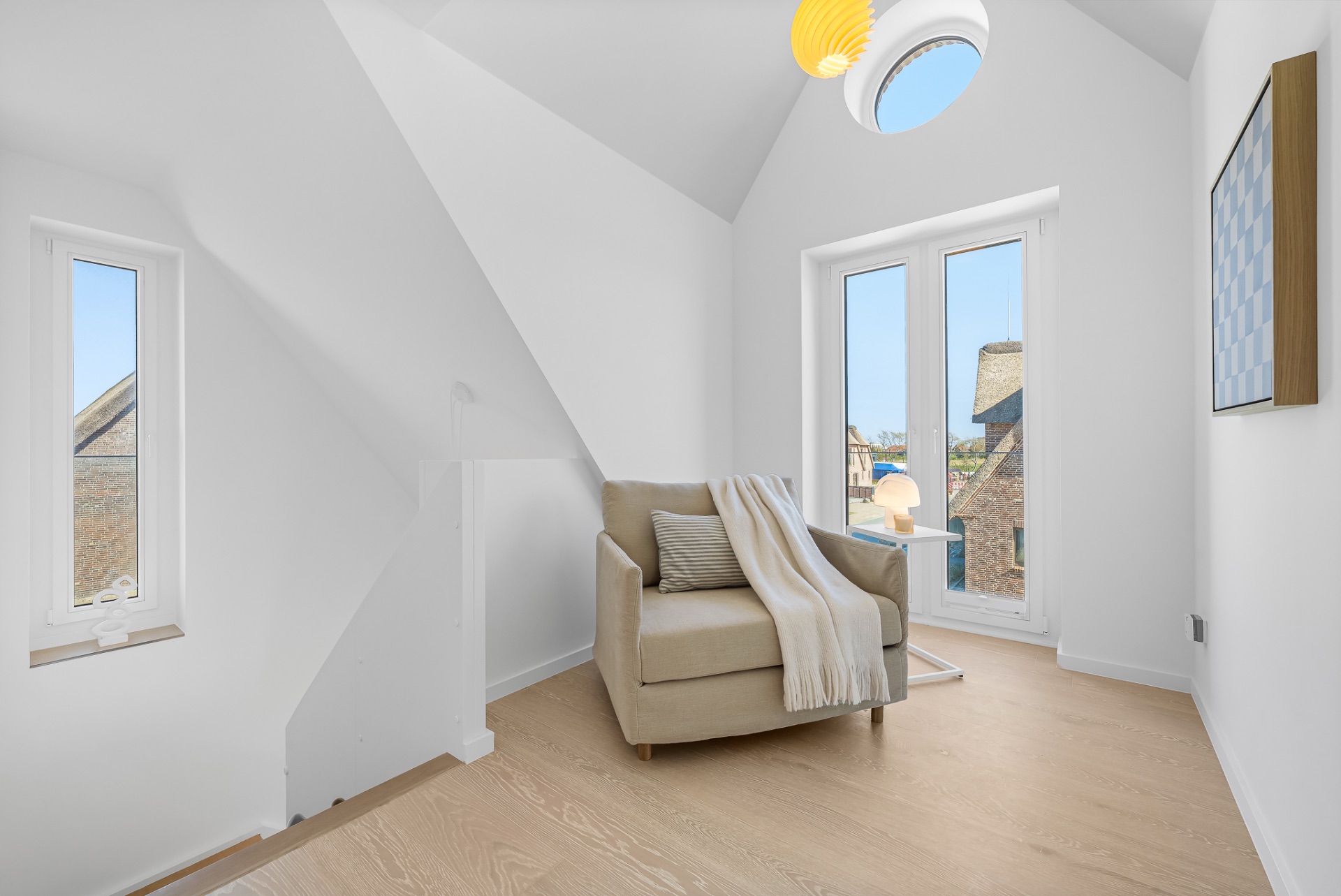 Helles Zimmer mit Sessel, Kissen, Decke, zwei Fenstern und Zugang zu einem kleinen Balkon.