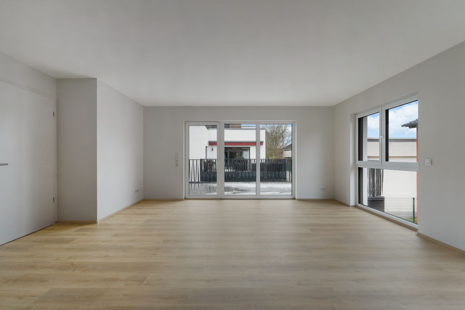 Leerer, heller Raum mit Holzfußboden und großen Fenstern, Blick auf eine Terrasse.
