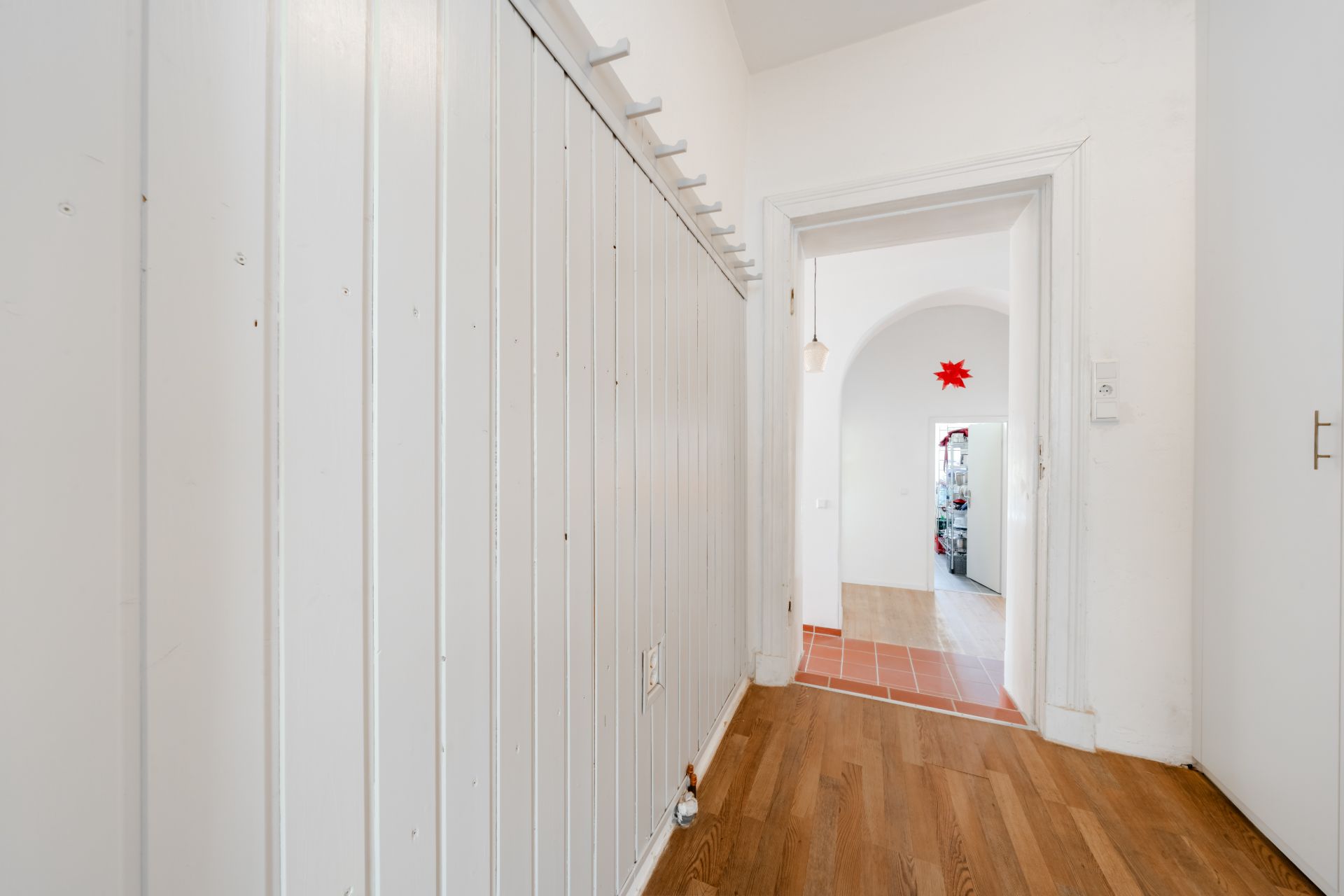 Flur mit Holzfußboden und weißer Wand, Blick durch offene Tür in weiteren Raum.