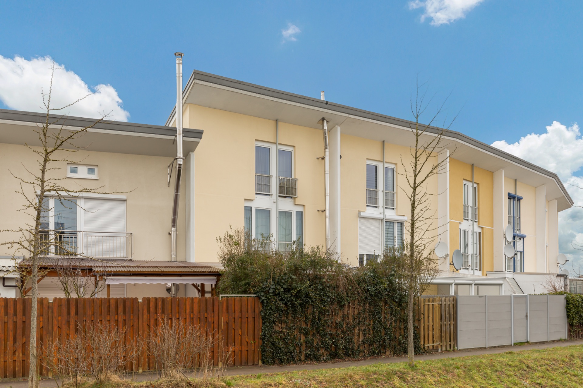 Mehrfamilienhaus mit gelber Fassade, Holzzaun und kleinen Bäumen davor.