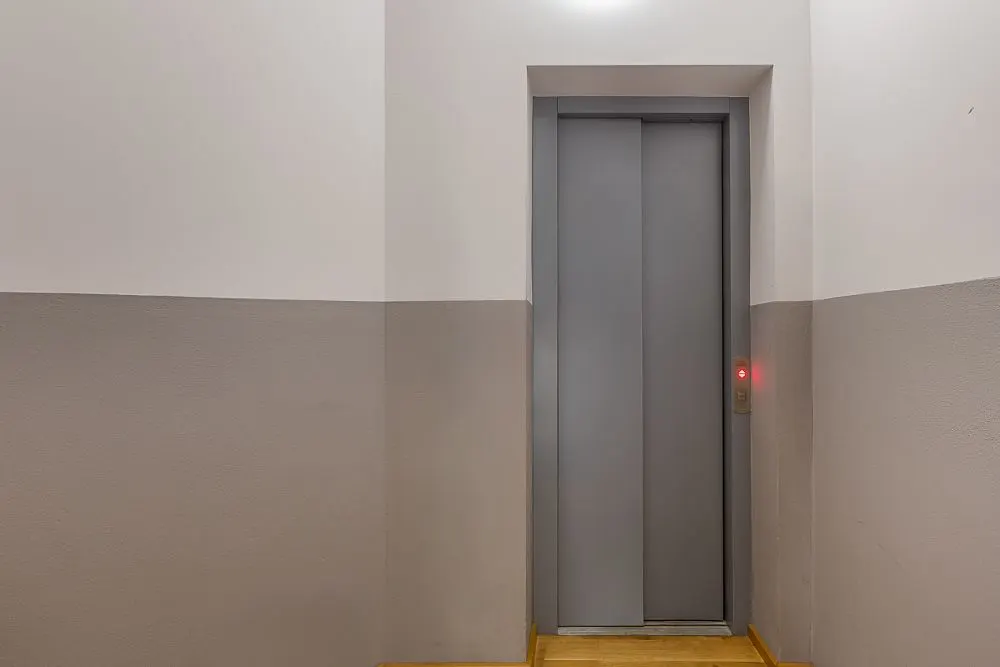Geschlossener Aufzug mit grauen Türen und einem beleuchteten Knopf an der Wand.