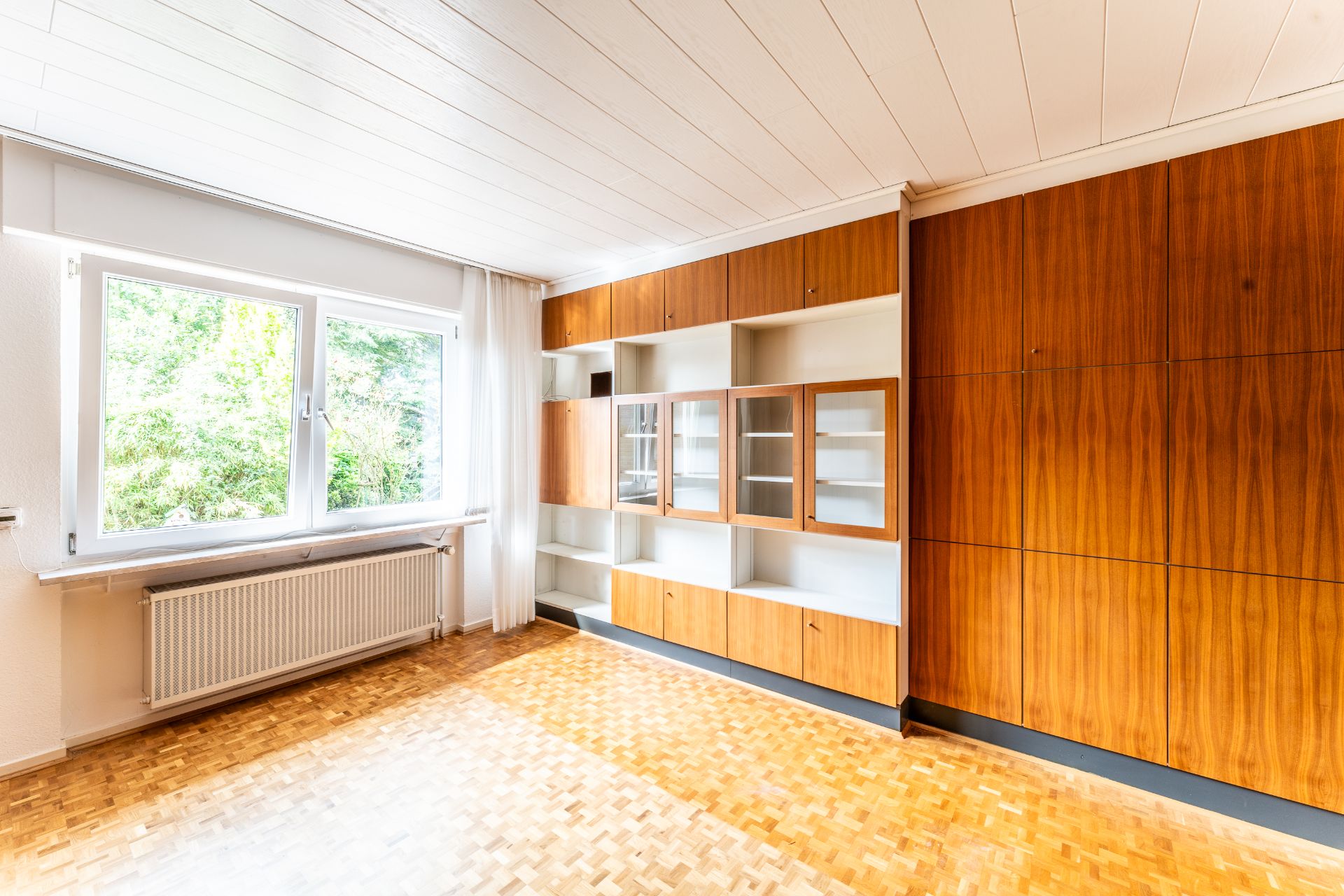 Helles Zimmer mit großem Fenster, Parkettboden und Einbauschränken aus Holz.