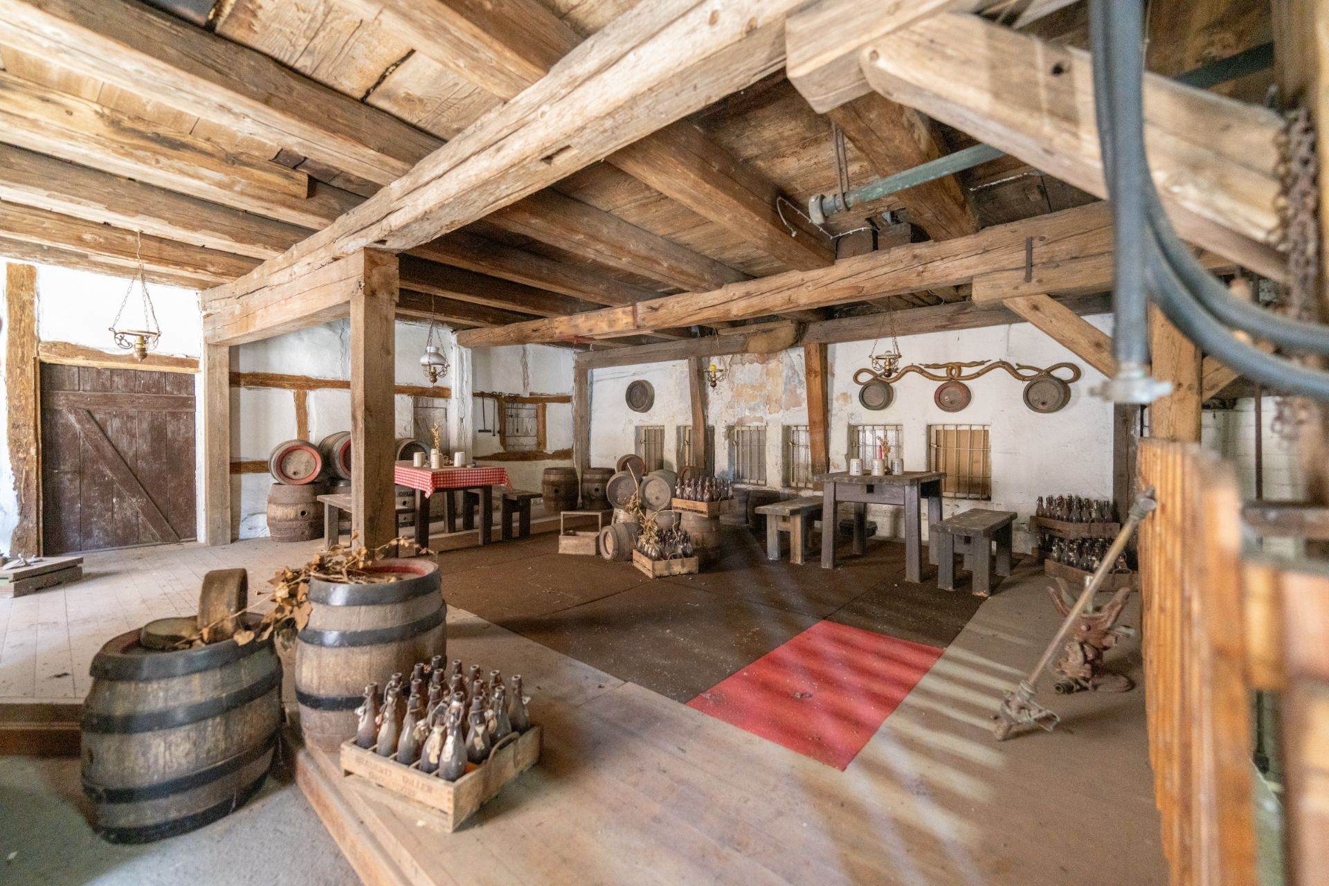 Rustikaler Raum mit Holzbalken, Fässern, Tischen und Bänken, dekoriert mit alten Flaschen.