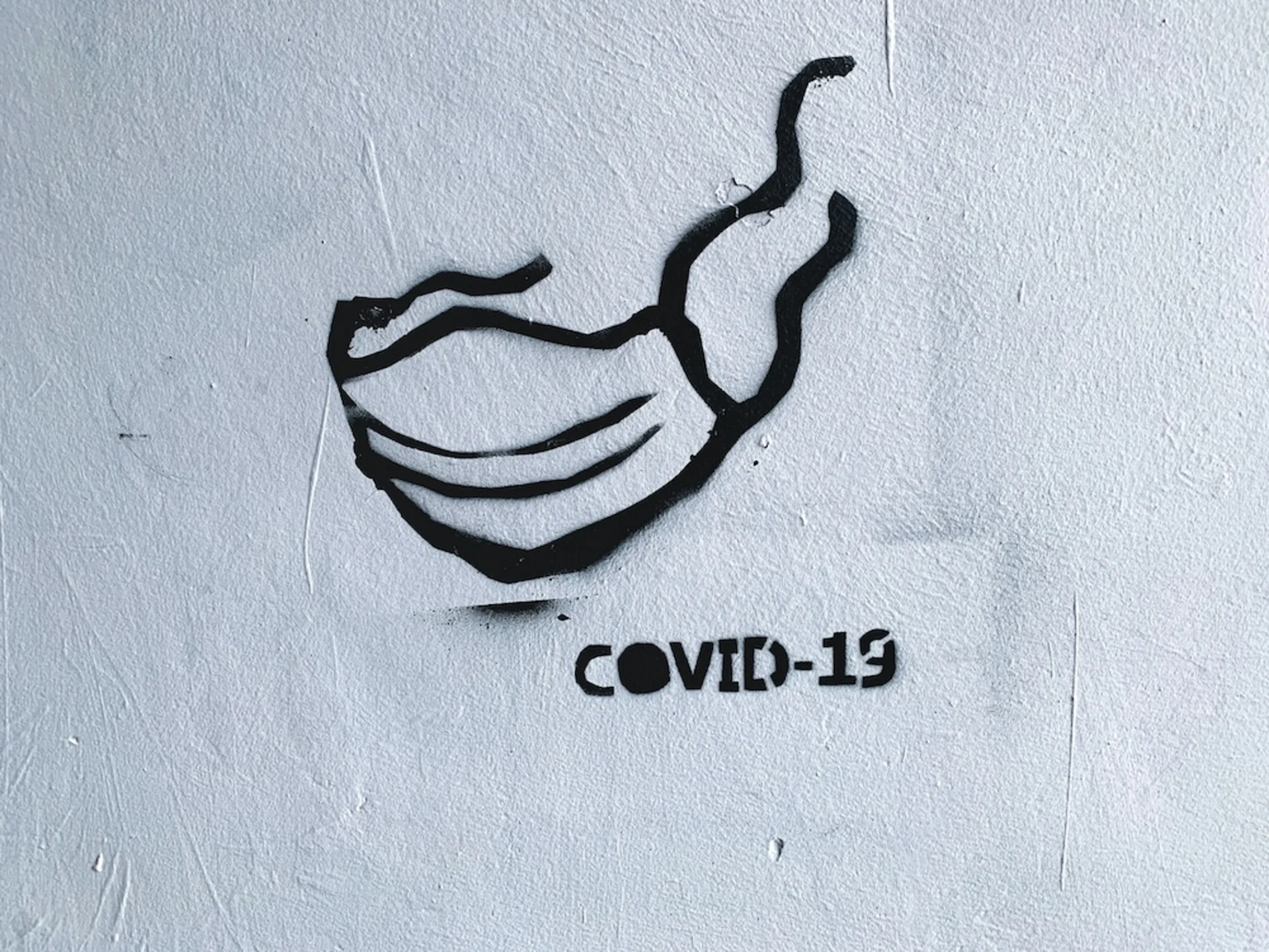 Schwarze Zeichnung einer Maske und der Schriftzug 'COVID-19' auf einer weißen Wand.