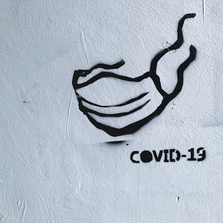 Schwarze Zeichnung einer Maske und der Schriftzug 'COVID-19' auf einer weißen Wand.