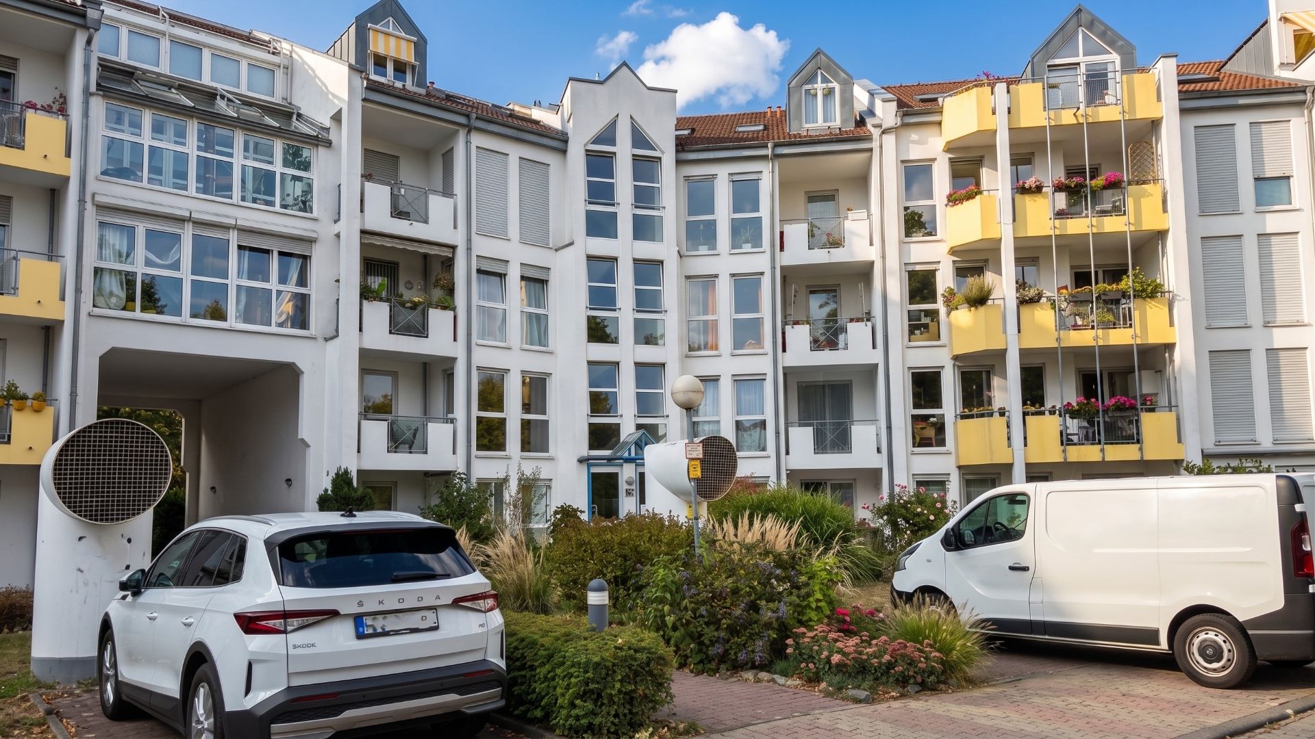 Mehrstöckiges Wohnhaus mit Balkonen, geparktem Auto und Van davor, umgeben von Pflanzen.