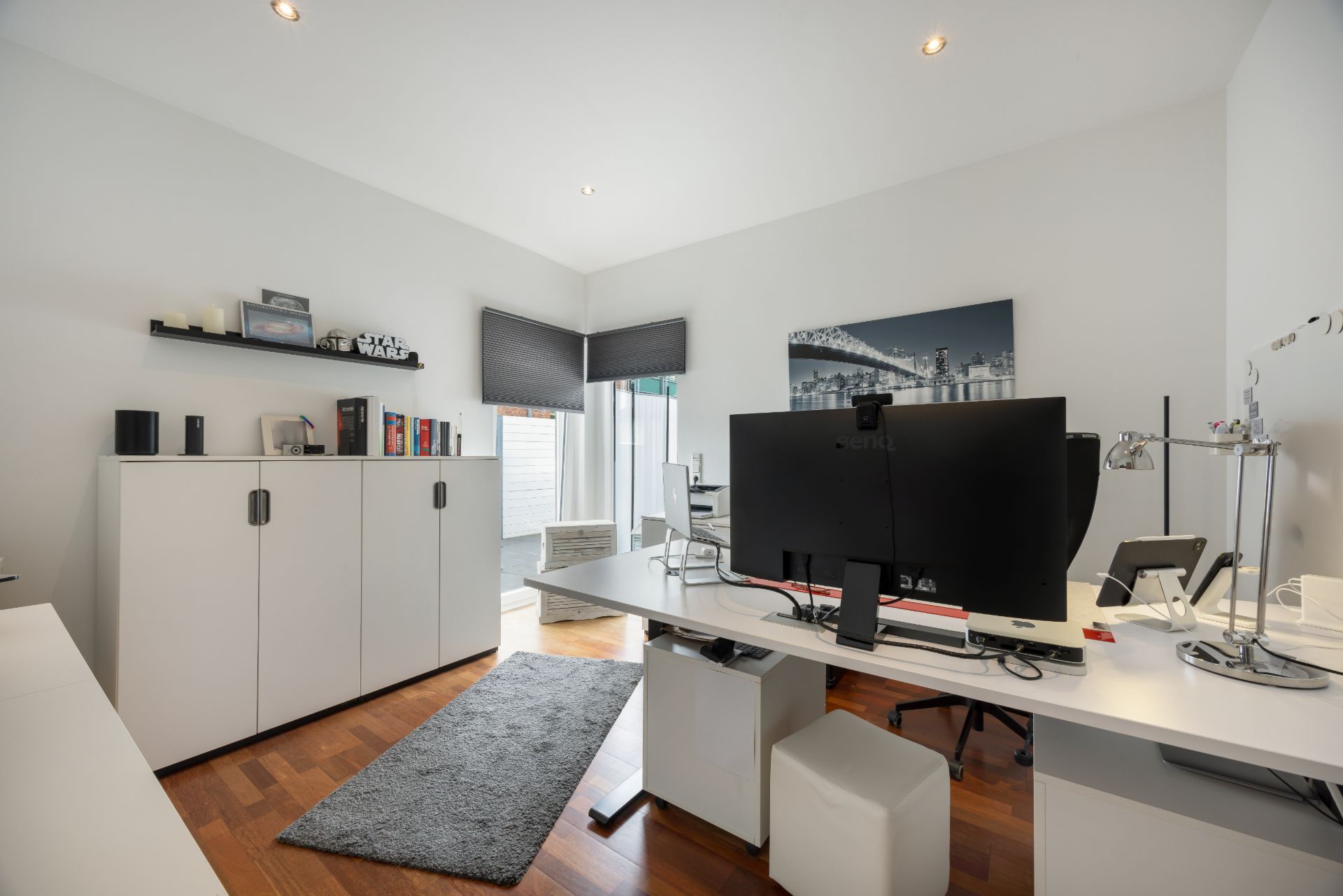 Modernes Arbeitszimmer mit großem Schreibtisch, Computer, Regalen und weißem Schrank auf Holzboden.