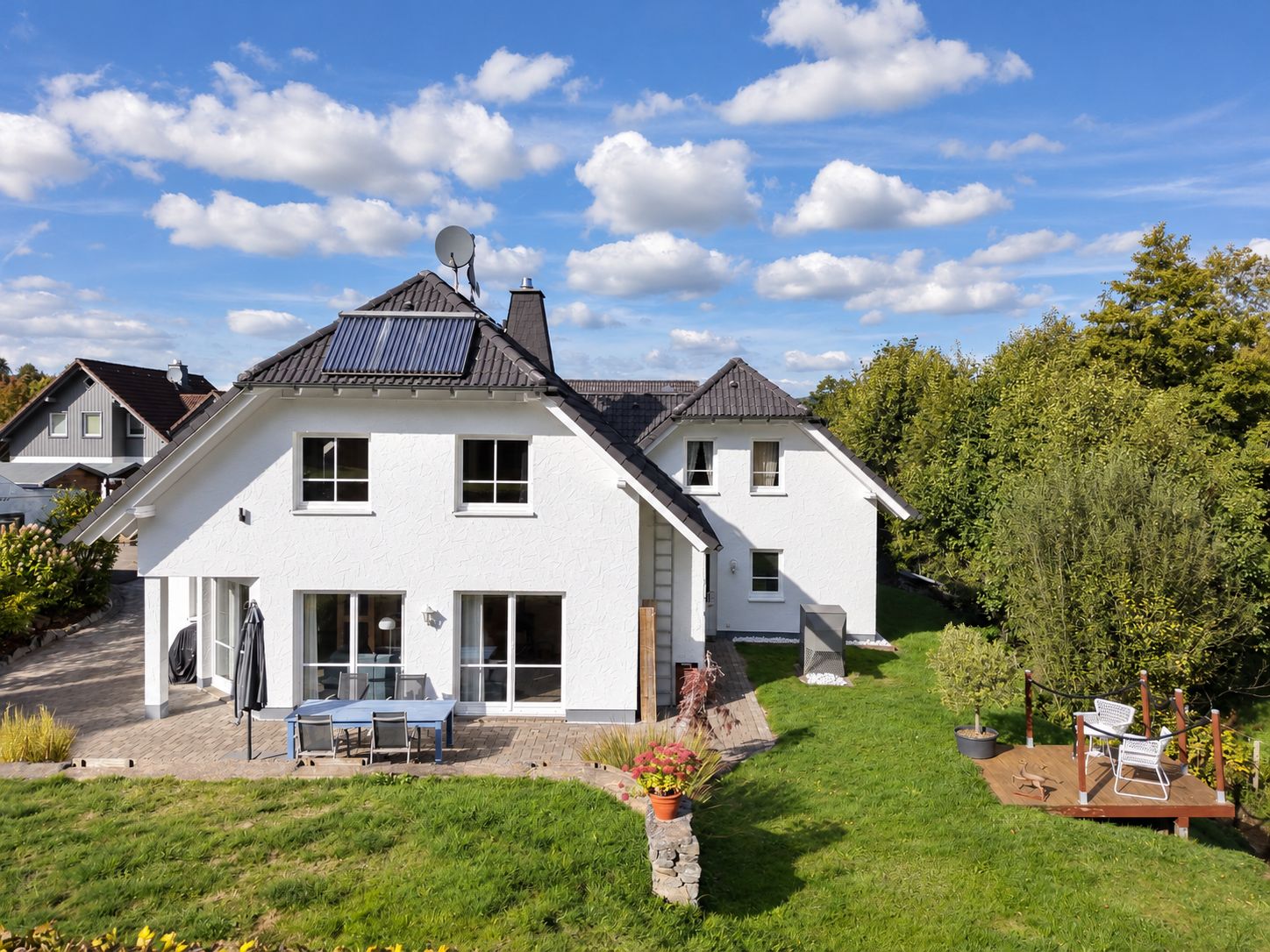 Weißes Einfamilienhaus mit Garten, Terrasse und Gartenmöbeln bei sonnigem Wetter.