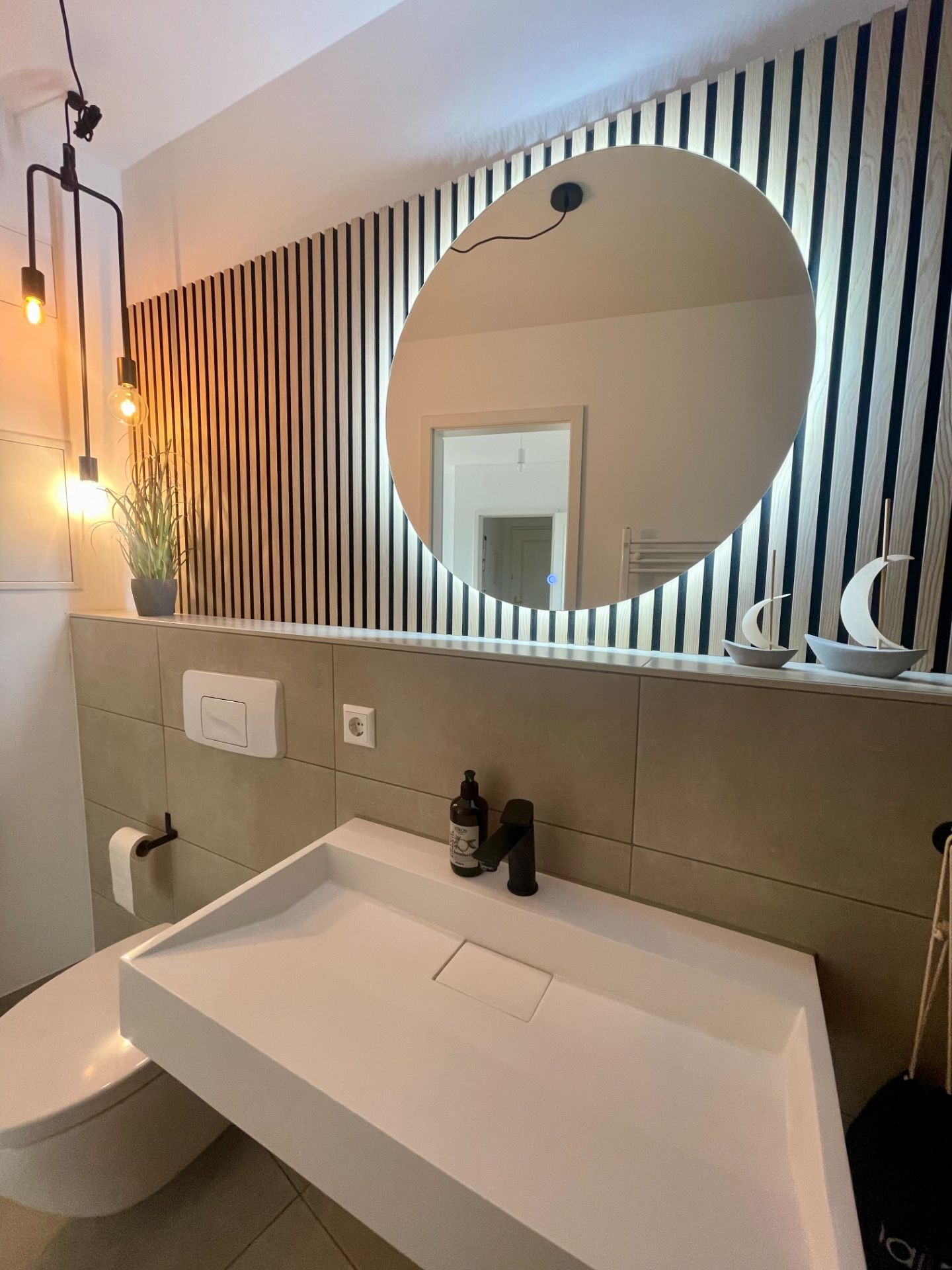 Modernes Badezimmer mit großem rundem Spiegel, Waschbecken, WC und dekorativen Lampen.