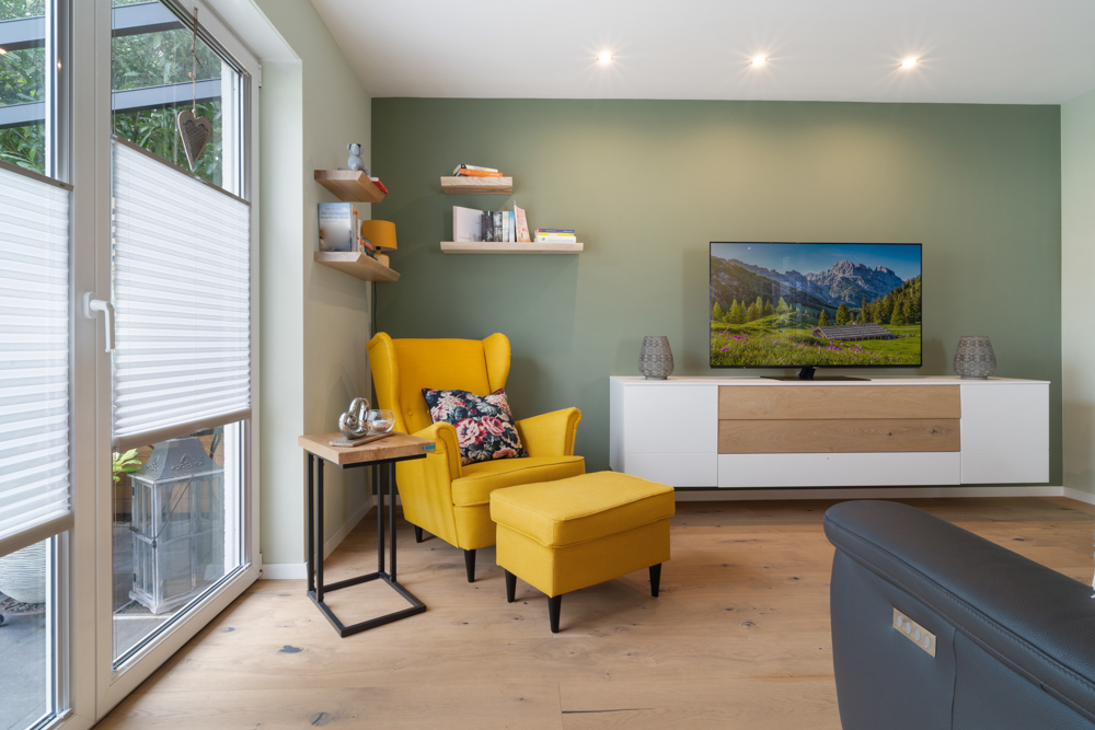 Modernes Wohnzimmer mit gelbem Sessel, Beistelltisch, TV und Regal an grüner Wand.