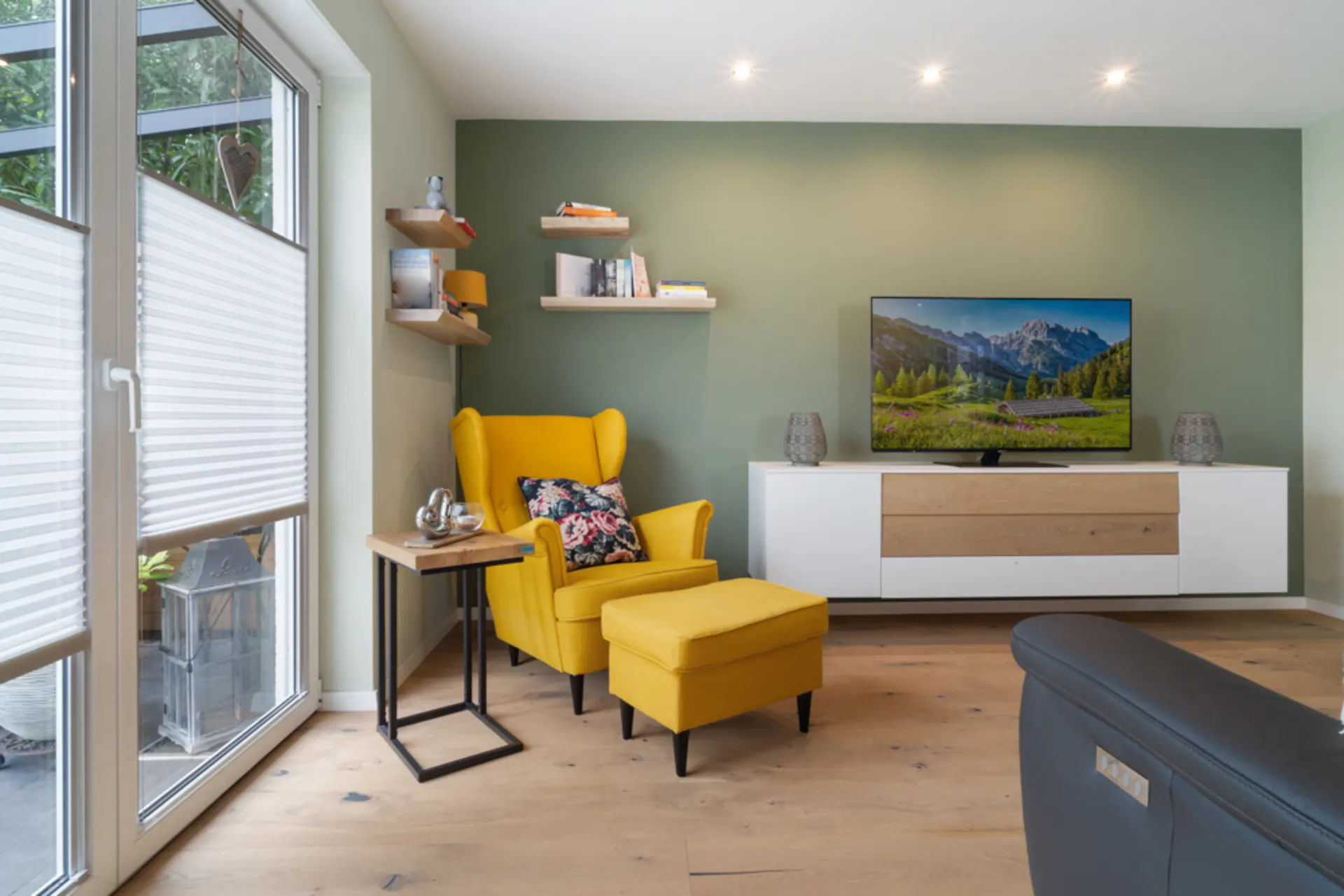 Modernes Wohnzimmer mit gelbem Sessel, Beistelltisch, TV und Regal an grüner Wand.
