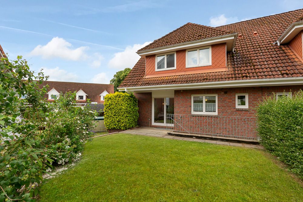 Backsteinwohnhaus mit rotem Dach, Vorgarten und Terrasse, umgeben von Büschen und Rasen.