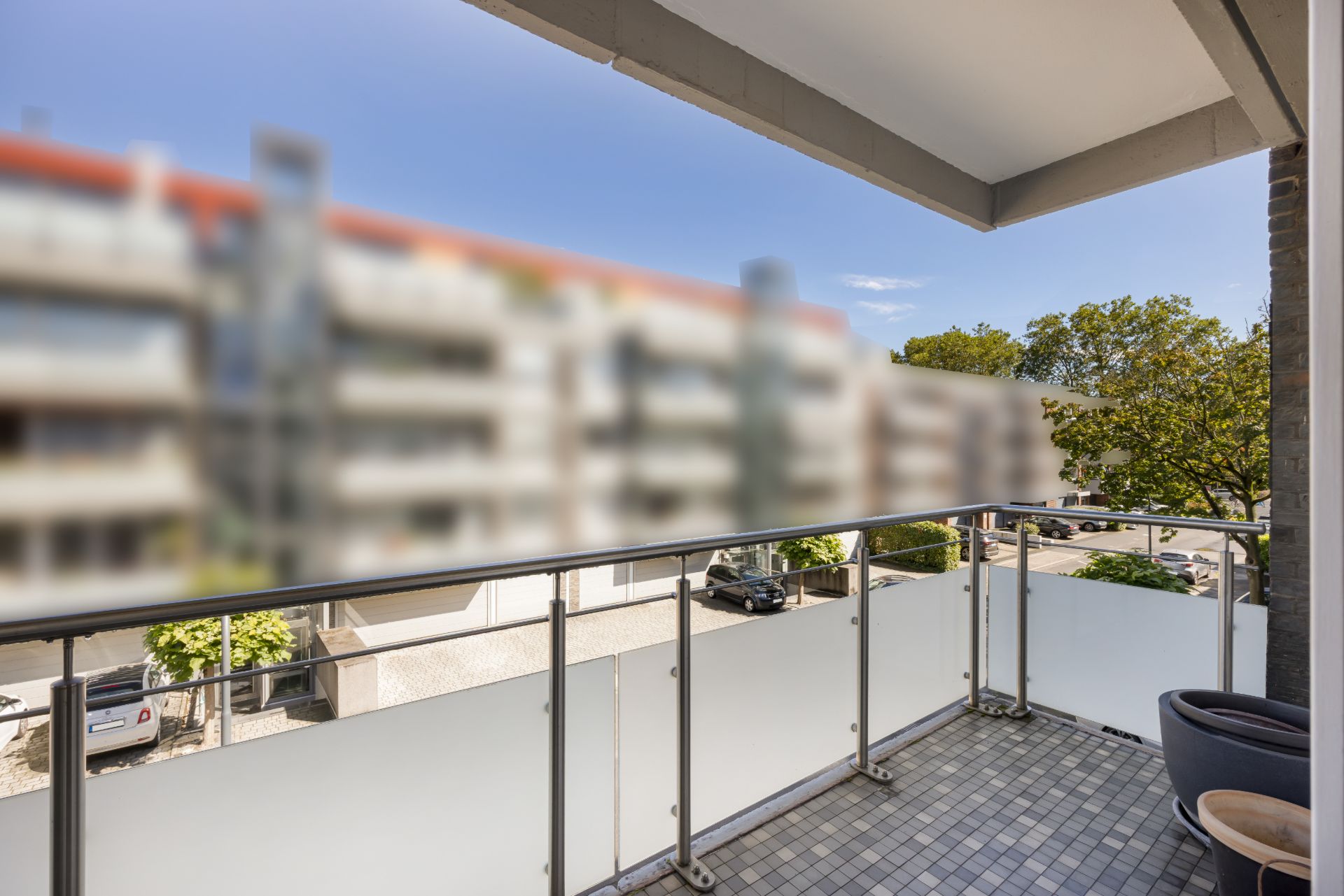 Balkon mit Glasgeländer, Blick auf Straße und Bäume, zwei Blumentöpfe in der Ecke.