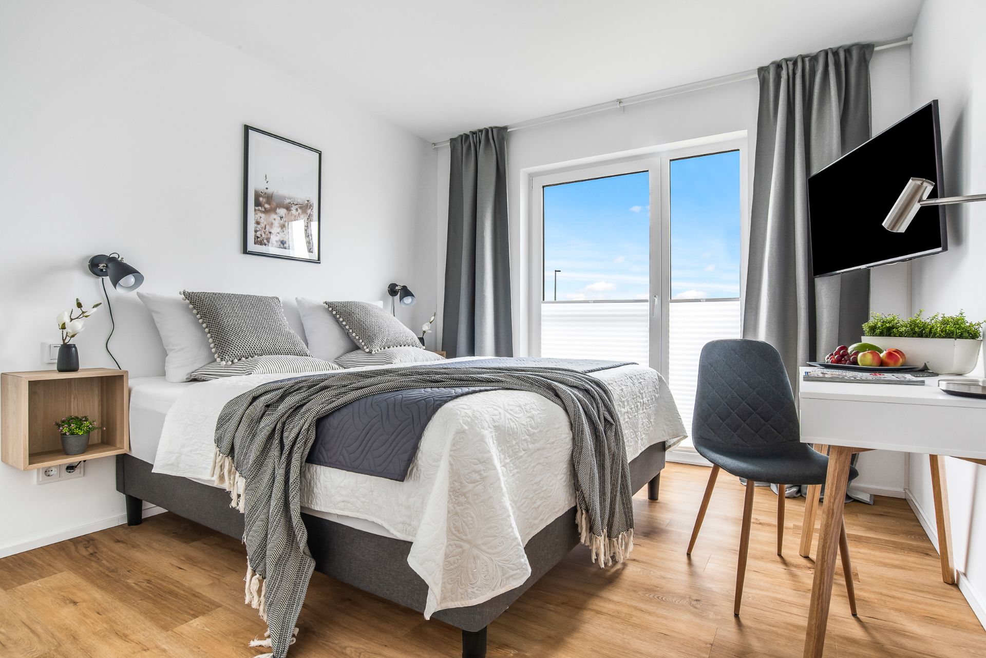 Helles Schlafzimmer mit Doppelbett, Schreibtisch, Stuhl und großem Fenster mit grauen Vorhängen.