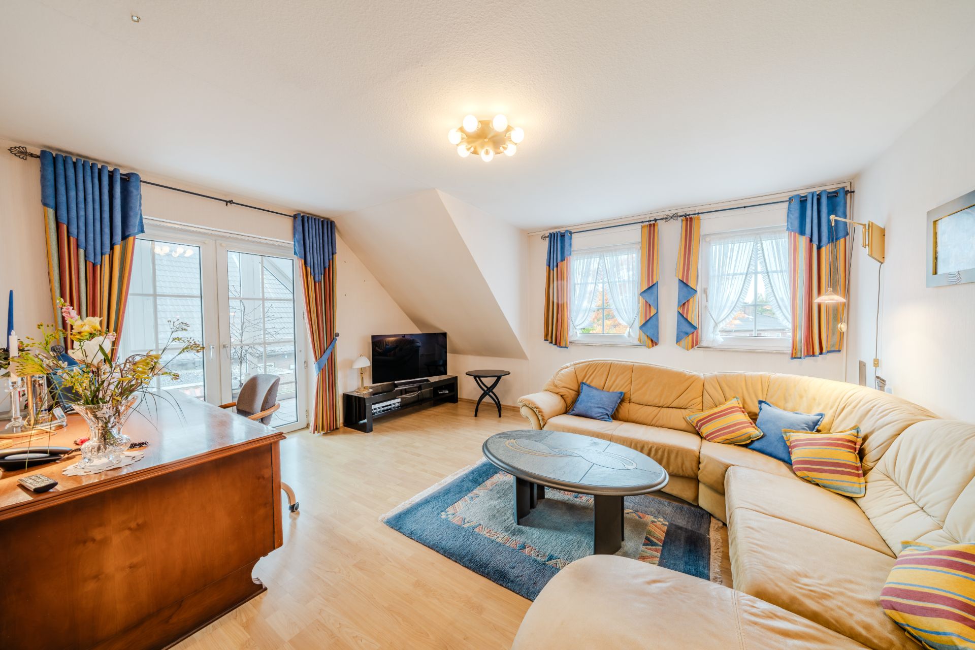 Helles Wohnzimmer mit großer Eckcouch, Couchtisch, TV und mehreren Fenstern mit bunten Vorhängen.
