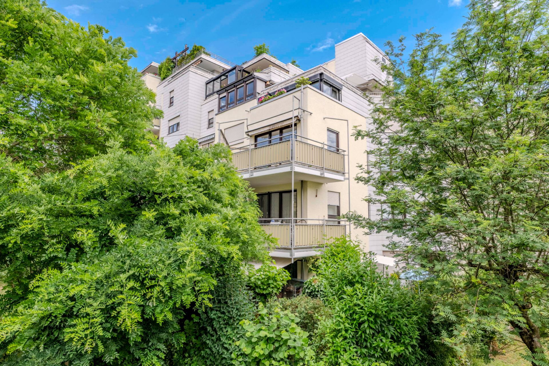 Mehrstöckiges Wohnhaus mit Balkonen, umgeben von vielen grünen Bäumen und Pflanzen.