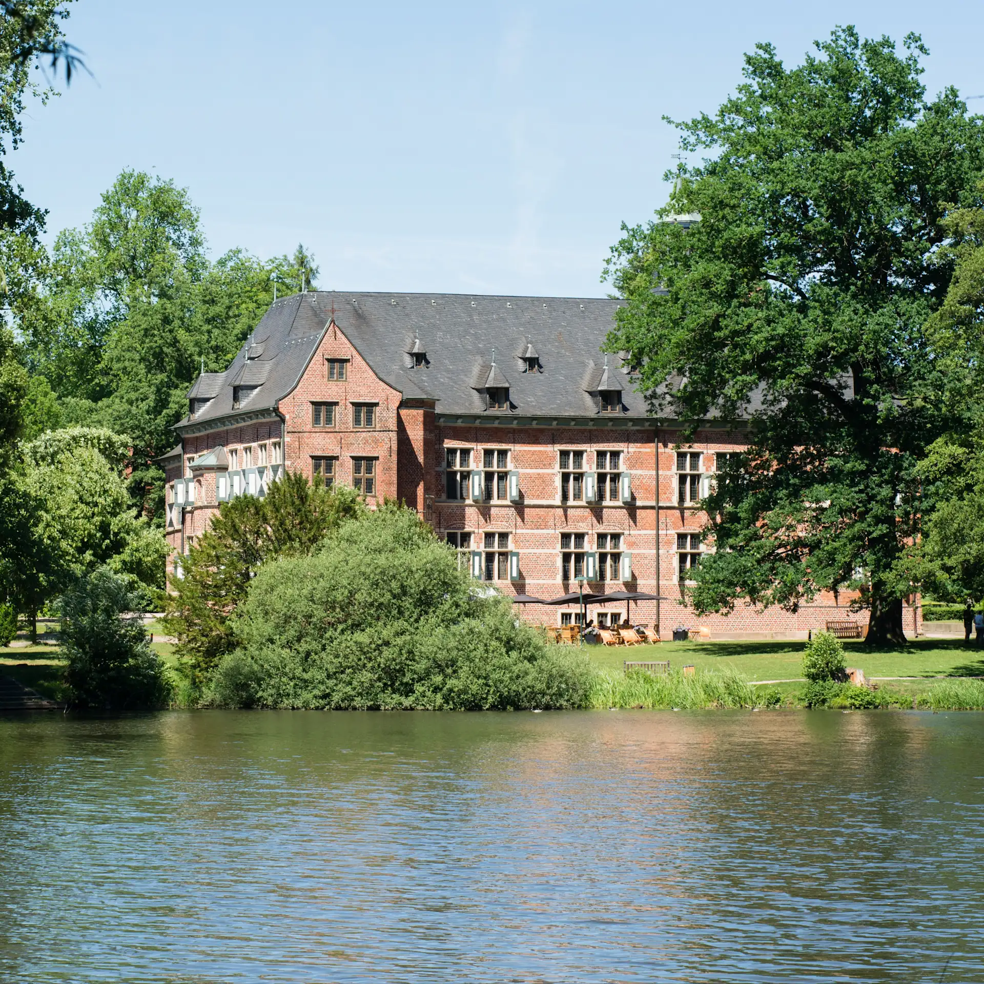 Großes Backsteingebäude mit vielen Fenstern, umgeben von Bäumen und einem Teich im Vordergrund.
