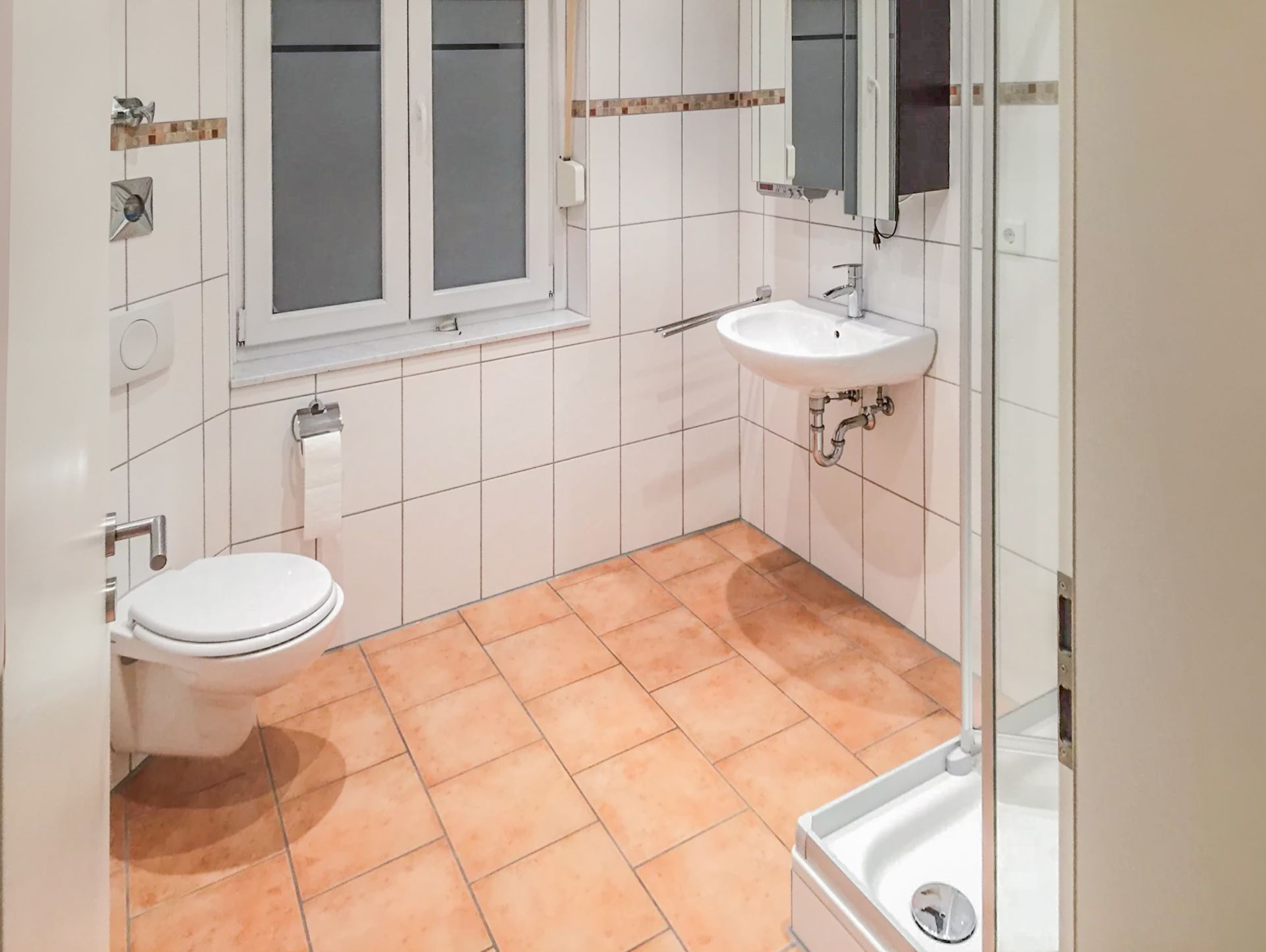 Helles Badezimmer mit Toilette, Waschbecken, Dusche, Fenster und Fliesenboden.