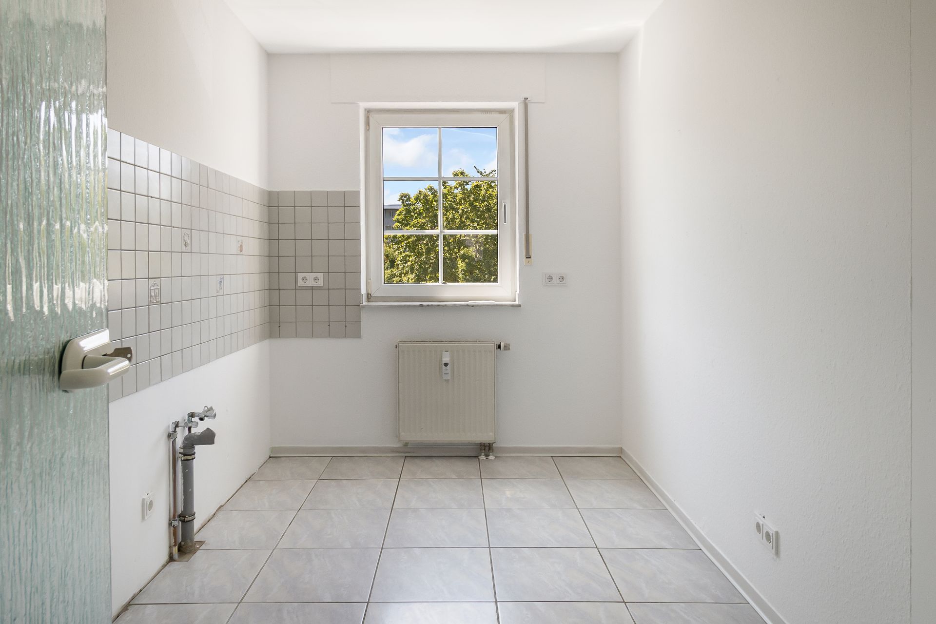 Heller, leerer Raum mit Fliesenboden, Fenster und Heizkörper, weiße Wände und geflieste Ecke.