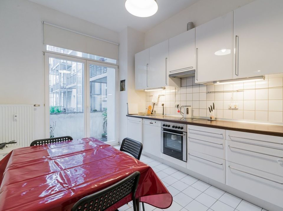 Moderne weiße Küche mit Esstisch, roten Tischdecke, großen Fenstern und viel Tageslicht.